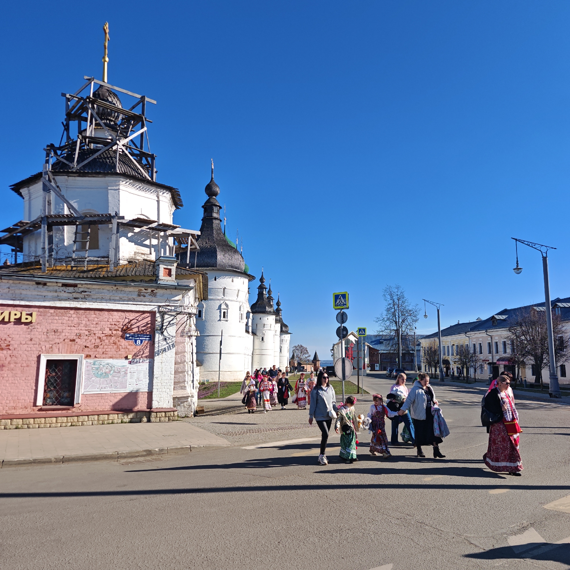The image captures a vibrant street scene in Rostov, Russia, under a clear blue sky. A group of people, including adults and children, are crossing an asphalt road on a sunny day, indicated by the bright light and long shadows. Many individuals are dressed in traditional Russian folk costumes featuring intricate patterns, vibrant colors like red and white, and embroidered elements, suggesting a cultural event or celebration. Others are dressed in casual modern clothing.
To the left, a large white building with dark, conical cupolas and domes dominates the background. The uppermost dome, adorned with a golden cross, is surrounded by wooden scaffolding, indicating ongoing construction or renovation work. Adjacent to this main structure, a pink brick building with white trim and a small barred window is visible.
Several street elements are present: a pedestrian crossing sign, a yellow and black "pedestrian ahead" road sign, and ornate black streetlights lining the road. In the distance, more buildings with similar architectural styles can be seen, along with bare trees and parked cars.
Visible text includes:
- On the pink brick building, partial Cyrillic text "ИРЬ" is visible.
- A blue street sign on the pink building reads "УЛИЦА 50-ЛЕТИЯ ВЛКСМ" (Street of the 50th Anniversary of Komsomol) with the number "8" below it.
- A map displayed on the wall of the pink building contains Cyrillic text, including "АКУН".
The scene evokes a sense of local culture and history, with the traditional costumes contrasting with modern elements, all set against the backdrop of historic Russian architecture.
