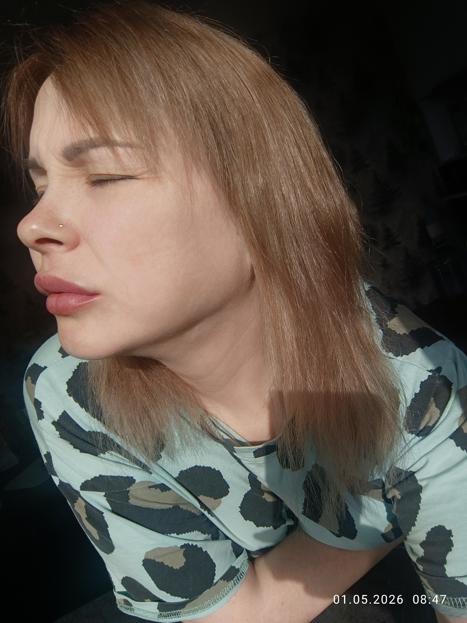 The image displays a close-up, side profile of a woman with light brown hair, seen from her chest upwards. She is looking to the left, with her eyes squinted or closed, likely due to bright sunlight illuminating her face and hair from the right side of the frame. Her lips are slightly parted. She has a small, gold stud nose piercing on her right nostril. She is wearing a light greenish-blue t-shirt adorned with a dark, abstract, leopard-like print. The background is dark and out of focus, suggesting an indoor setting. A digital timestamp, "01.05.2026 08:47", is visible in the bottom right corner of the original image, indicating the date and time of capture. The strong, direct light suggests a sunny morning. The location context provided is Chaikovsky, Russia.