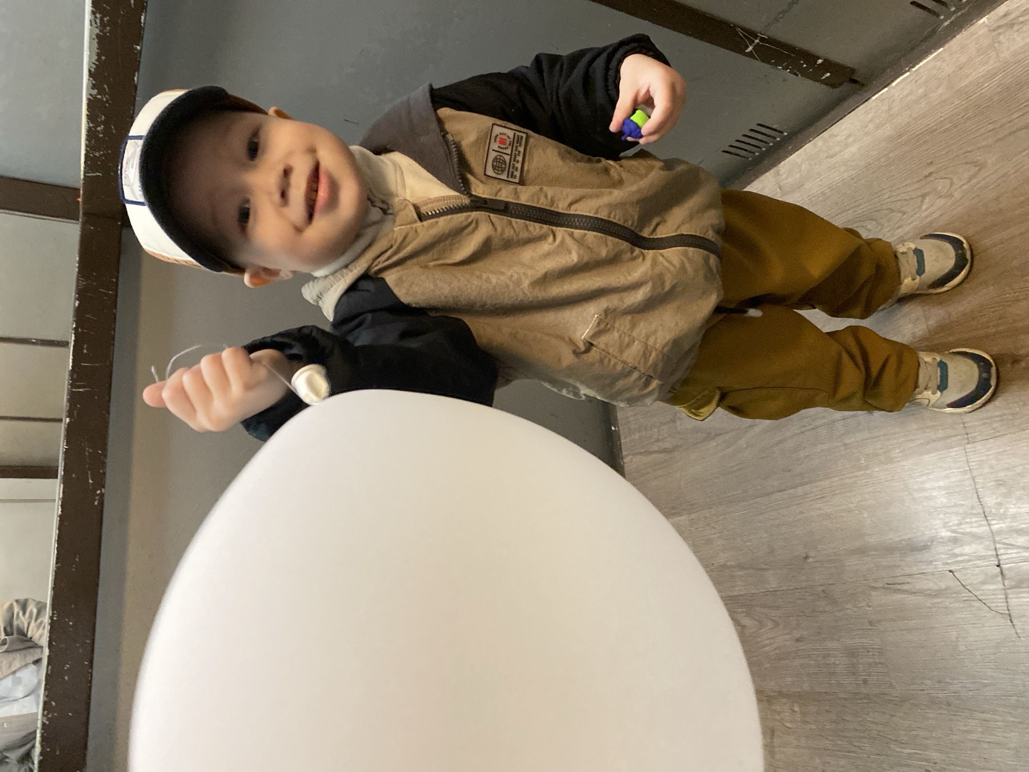 A toddler stands indoors with a large white balloon. The child wears a baseball cap, a light brown jacket with a black collar and sleeves, and mustard-colored pants. The child holds a small colorful toy in one hand and the string of the balloon in the other. The floor is wood-look laminate. A metal frame and cabinet are in the background. There is no evidence to support that this image was taken in Sochi, Russia.