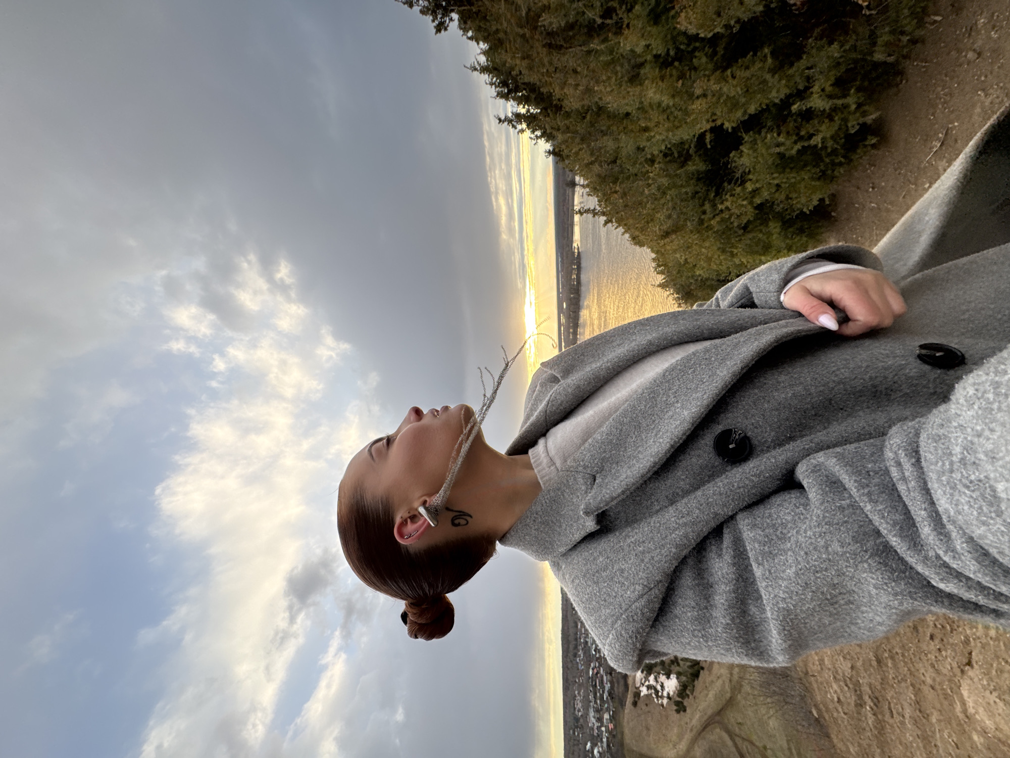 A serene outdoor scene captures a woman at what appears to be either sunrise or sunset, looking contemplatively towards the horizon. She is positioned on the right side of the frame, gazing upwards and to the left, with her profile visible. Her dark hair is neatly styled in a bun, and she wears a warm, grey woolen coat over a lighter-toned top. A prominent silver ear cuff and a delicate chain necklace are visible, along with a small, dark tattoo on her neck. Her right hand, with light-colored fingernails, rests near the buttons of her coat.
The background is dominated by a expansive sky filled with soft, cloudy formations, transitioning from bright, sunlit areas to cooler grey-blue tones. The sun, partially obscured by clouds, casts a warm, golden glow across a large body of water that stretches into the distance, reflecting the vibrant light. A line of verdant trees and dense foliage frames the top of the image, suggesting a natural, elevated setting. Below the illuminated water, a distant landscape hinting at a town or settlement can be seen. The scene evokes a tranquil and reflective mood, set in the natural environment of Olkhovka, Russia.