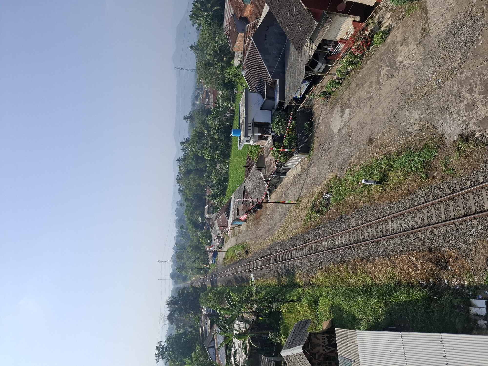 The image depicts a railway track running through a rural or suburban area, likely in Sukabumi, Indonesia, given the style of housing and vegetation. The sky is clear and pale blue, suggesting a bright, possibly warm day. A double set of train tracks dominates the left side of the frame, stretching into the distance. The tracks are laid on ballast, with sparse vegetation growing along the sides.
To the right of the tracks, a dirt road or pathway runs parallel. Along this road, several modest buildings with tiled roofs are visible. These structures appear to be houses or small shops, some adorned with decorative flags or banners, possibly indicating a local celebration or event. The buildings feature a mix of architectural styles, from simple single-story structures to a more modern two-story building with a blue water tank on its roof. Some buildings have open-fronted sections, possibly serving as workshops or storefronts. Fences and vegetation surround the properties.
In the foreground on the left, there is a corrugated metal structure with the word "ABADI" visible on its side. Further back, the landscape is filled with lush green trees and vegetation, typical of a tropical environment. In the far distance, rolling hills or mountains can be seen, partially obscured by a light haze. Electrical power lines and transmission towers are also visible in the middle and far distance, indicating the presence of infrastructure. The scene is devoid of any visible people or vehicles actively engaged in activity. The overall impression is one of a quiet, everyday scene in a developing community.