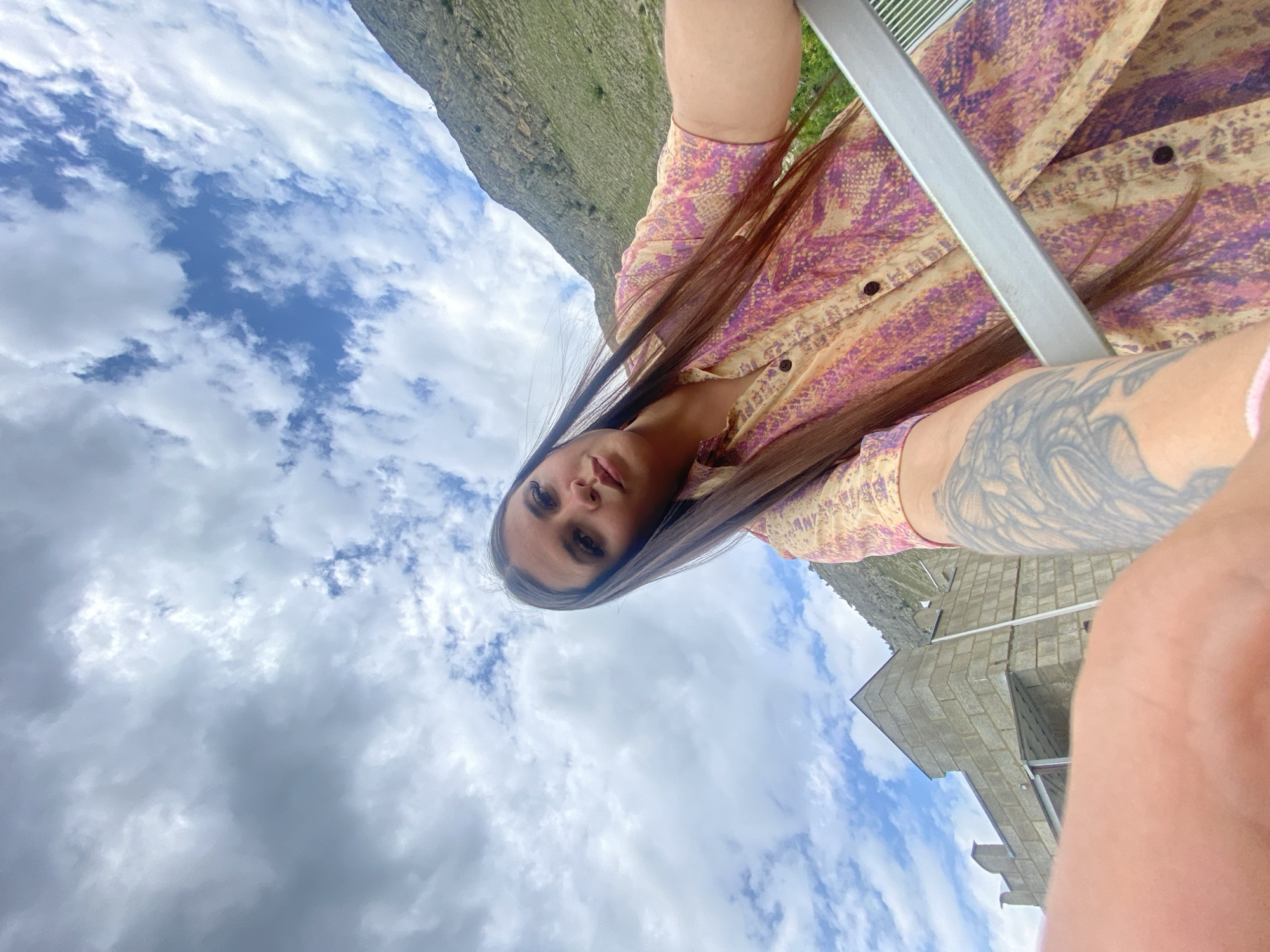 A young woman with long, dark hair and makeup is captured in a low-angle selfie. She is wearing a short-sleeved, collared shirt with a snakeskin-like pattern in shades of pink, purple, and yellow. A tattoo is visible on her left arm. She is outdoors, with a cloudy sky above. To the left, a stone building with a triangular roof and decorative turrets is visible, possibly a structure in the city of Chalda, Russia. In the background, a grassy, mountainous landscape rises up. A silver metallic object, possibly part of a railing or furniture, runs across the foreground of the image.