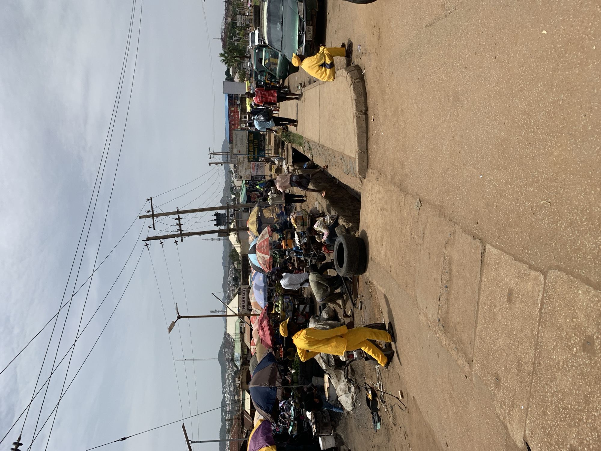 The image shows a street scene in Rafin Par, Nigeria. The view is from an elevated perspective, looking down onto a busy sidewalk and street. People are walking along the sidewalk and browsing goods displayed under umbrellas. A tire is leaning against some of the goods. Two people wearing yellow outfits are visible on the edge of the sidewalk. Power lines crisscross above the street. In the distance, there are buildings and hills. A vehicle and more people are further up the road. The sky is overcast.