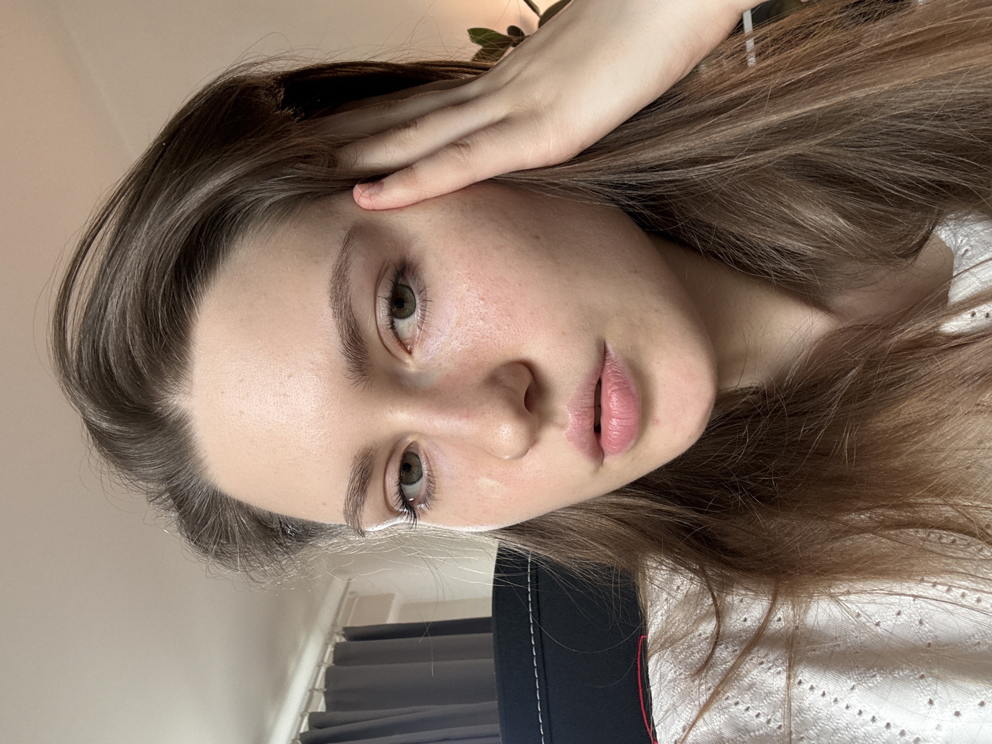 A close-up portrait of a young woman with light brown hair and fair skin. Her eyes are light green. She is looking at the camera, with her hand resting on her forehead. Her hair is parted on the side and falls around her face. She has light makeup.
The background is blurred, but there is a glimpse of a white wall and a plant. The setting does not allow to determine its location in Saint Petersburg, Russia.