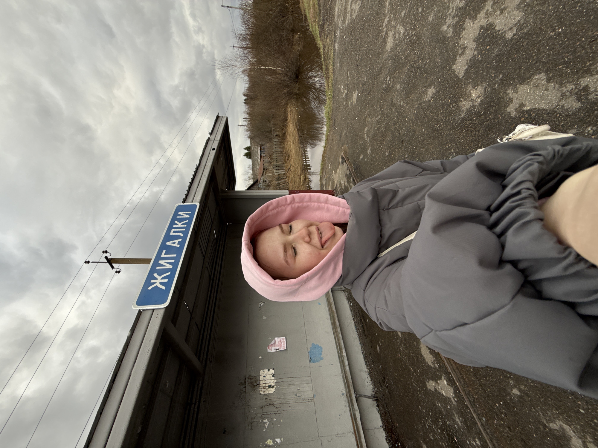 The image, oriented vertically, captures a playful moment featuring a young individual in Zhigalki, Russia. The person, seen from the chest up, wears a grey puffer jacket with a pink hoodie pulled over their head. Their eyes are closed, and their tongue is slightly out, conveying a lighthearted and relaxed expression.
They are positioned next to a simple, roofed shelter, likely a bus stop or small station. A prominent blue sign with white Cyrillic text, clearly spelling "ЖИГАЛКИ" (ZHIGALKI), is mounted on a pole beside this structure, confirming the location. Power lines extend across the frame, connected to a wooden utility pole visible in the mid-ground.
The outdoor setting appears rural or semi-rural under an overcast sky filled with grey clouds. The paved ground in the foreground is damp in places, suggesting recent rain or wet conditions. Beyond the immediate structure, the background features dense, bare bushes and trees, characteristic of late autumn or early spring. Further in the distance, a wooden fence and a few old, weathered buildings are discernible, adding to the rustic atmosphere. The overall impression is a casual, everyday scene in a quiet, perhaps remote, location.