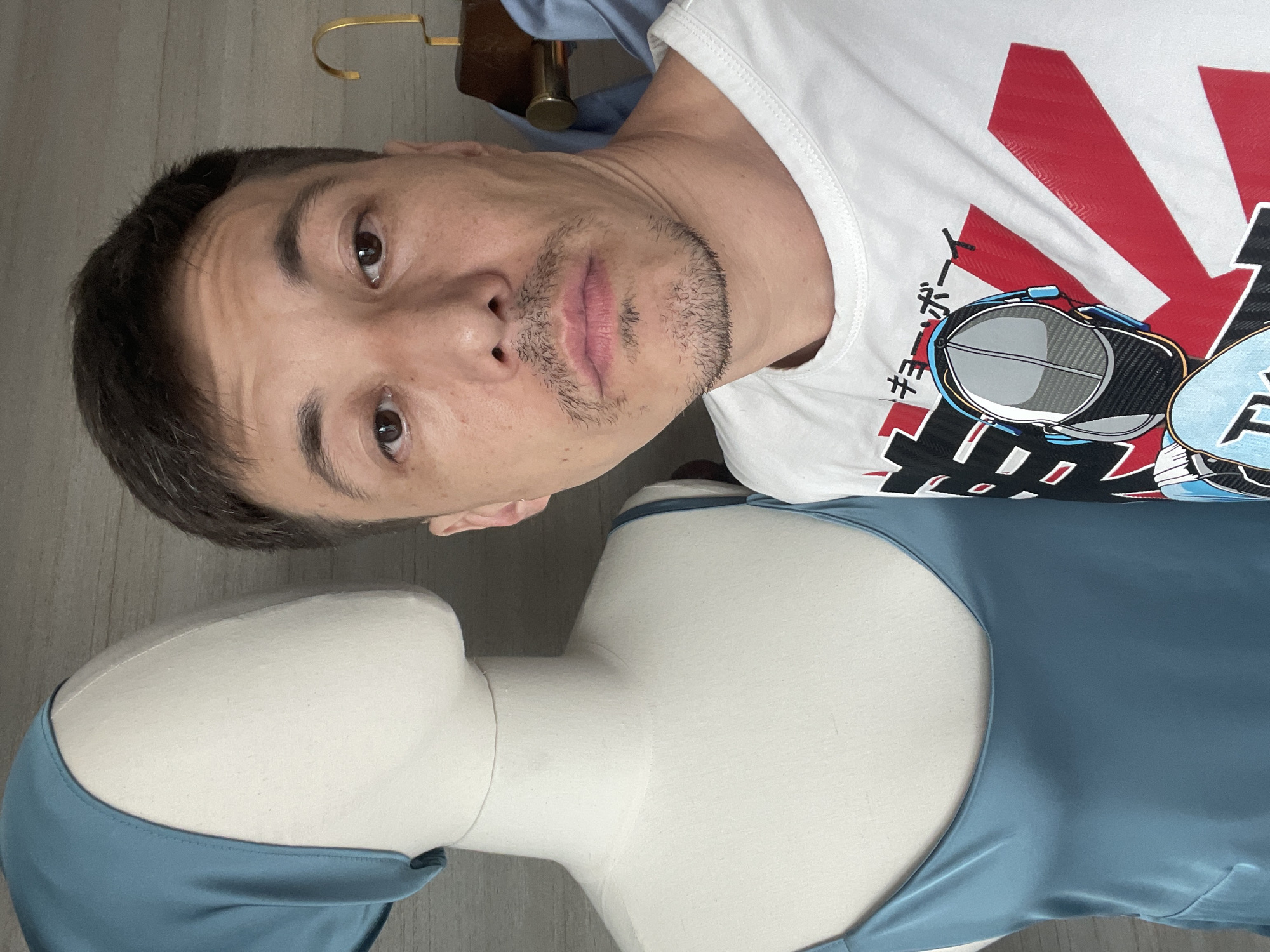 A man is positioned beside a mannequin wearing a blue dress. The location is implied to be Bac Nha Trang Ward, Vietnam. The man wears a white T-shirt with a red and black graphic design. The mannequin is featureless except for the blue dress, which covers its upper body. The background is a plain wall. The focus is on the man and the mannequin, with the environment blurred.