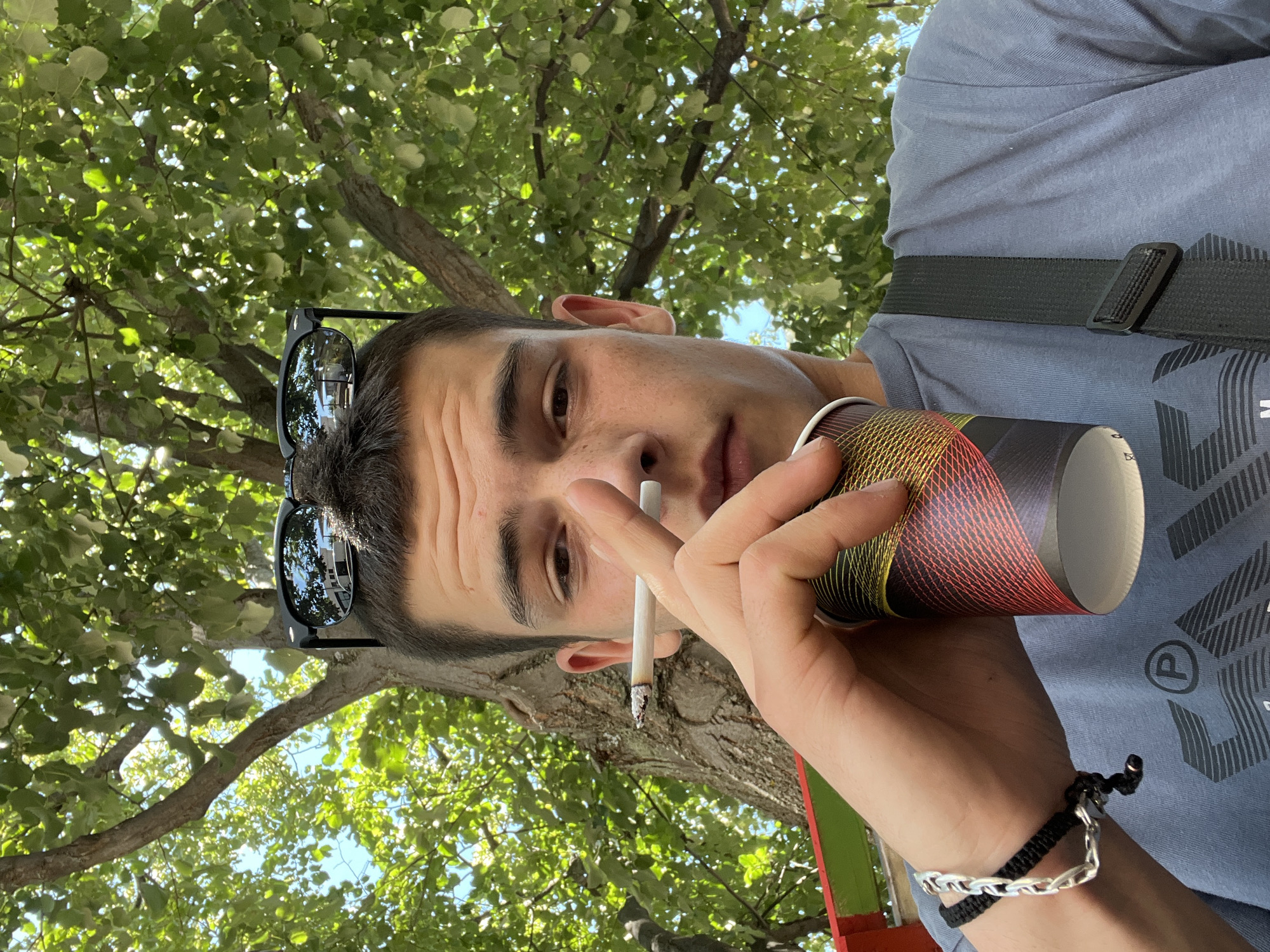 A young man with dark hair, wearing a blue t-shirt and black sunglasses perched on his head, holds a cigarette in his right hand and a colorful paper cup in his left. He is outdoors, with a large tree and lush green foliage behind him. The sun is shining, casting dappled light. The man appears to be looking directly at the camera. A black strap from a bag is visible crossing his chest. The overall setting suggests a casual outdoor moment. It is not possible to determine the precise location within the city of Sopot, Bulgaria from the provided image.