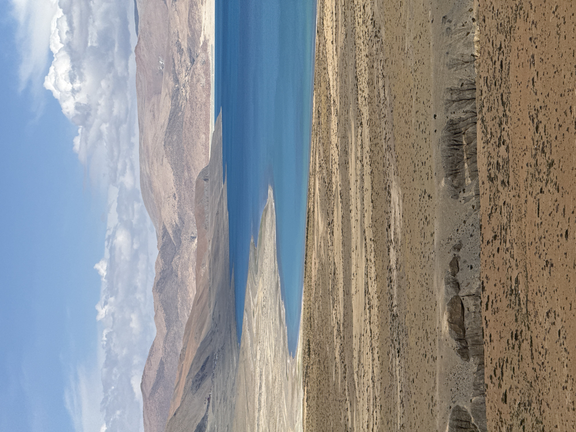 The image depicts a landscape in Gyirong County, China. The left side features a sky with scattered white clouds and a tan mountain range. In the center, a body of water is visible, with land extending into the water creating a peninsula. The right side is a dry, desert-like terrain with sparse vegetation. The scene appears to be taken from an elevated vantage point. There are no people visible in the scene.