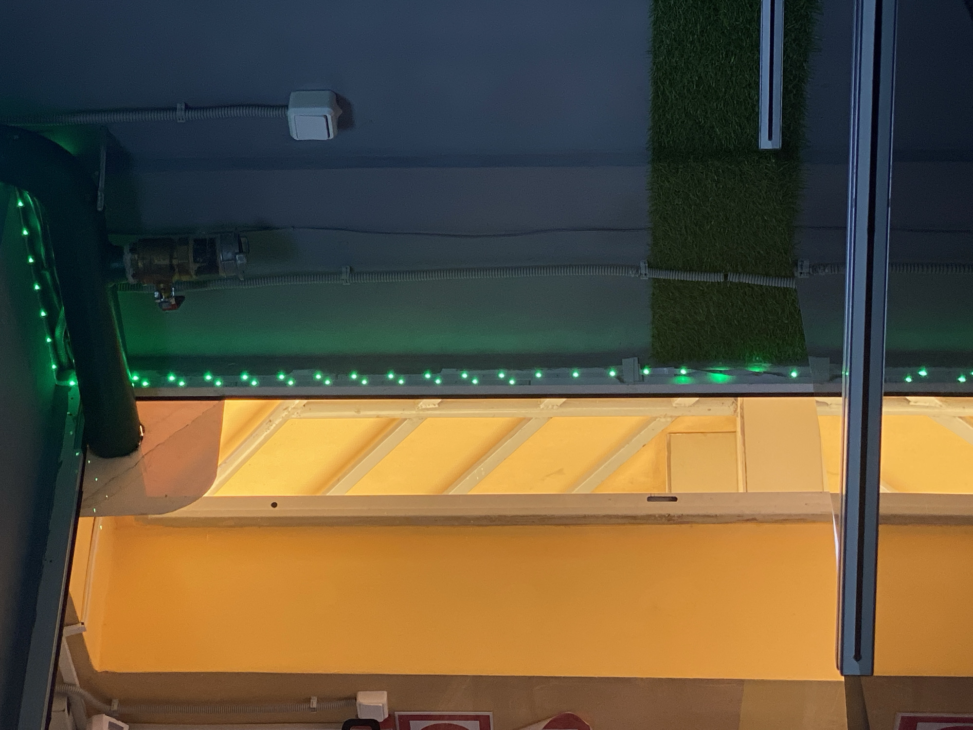 The image displays an architectural detail of what appears to be an indoor or sheltered outdoor space, possibly a service counter or storefront. The top section features a dark grey ceiling with various utilities: white electrical conduits, loose wires, and a white rectangular junction box or sensor. A dark green-colored pipe or column, decorated with a string of green LED lights, runs vertically down the left side. To its right, a vertical panel of artificial green turf is attached to the ceiling. Further right, a metallic strip or support structure is visible.
Below these overhead elements, another horizontal line of green LED string lights illuminates the top edge of a prominent yellow-orange structure. This structure appears to be an awning or canopy, with visible white or metallic support beams angled upwards underneath a lighter-colored, possibly fabric or panel, surface. Below this canopy, a large, brightly lit, uniform yellow-orange surface extends across the lower portion of the image, resembling a counter front or a wall. The artificial lighting, particularly the green LEDs and the bright yellow illumination, suggests an evening or nighttime setting, or an interior space. No people are visible, and no specific activity is taking place. Partially visible at the very bottom edge are the tops of red signs with white borders, but no text is legible.
