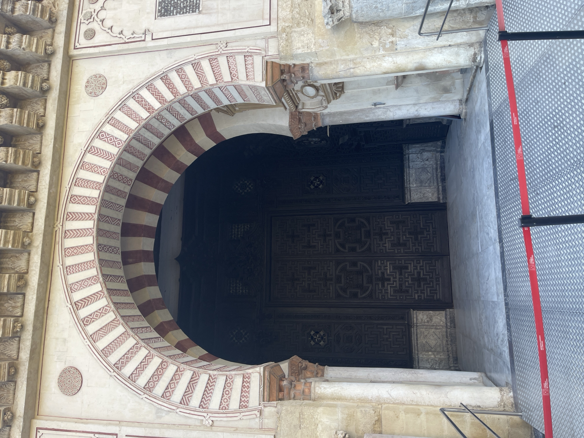 The image shows a large, ornate entranceway to the Mosque-Cathedral of Córdoba, Spain. The entrance features a horseshoe arch decorated with red and white stripes and geometric patterns. Flanking the entrance are columns and intricately carved stonework. The large, dark wooden doors are set back within the archway, and the area in front of them is paved with light-colored marble tiles. A red stanchion barrier with text in Spanish is in the foreground, indicating a restricted area. The overall impression is one of grandeur and historical significance.