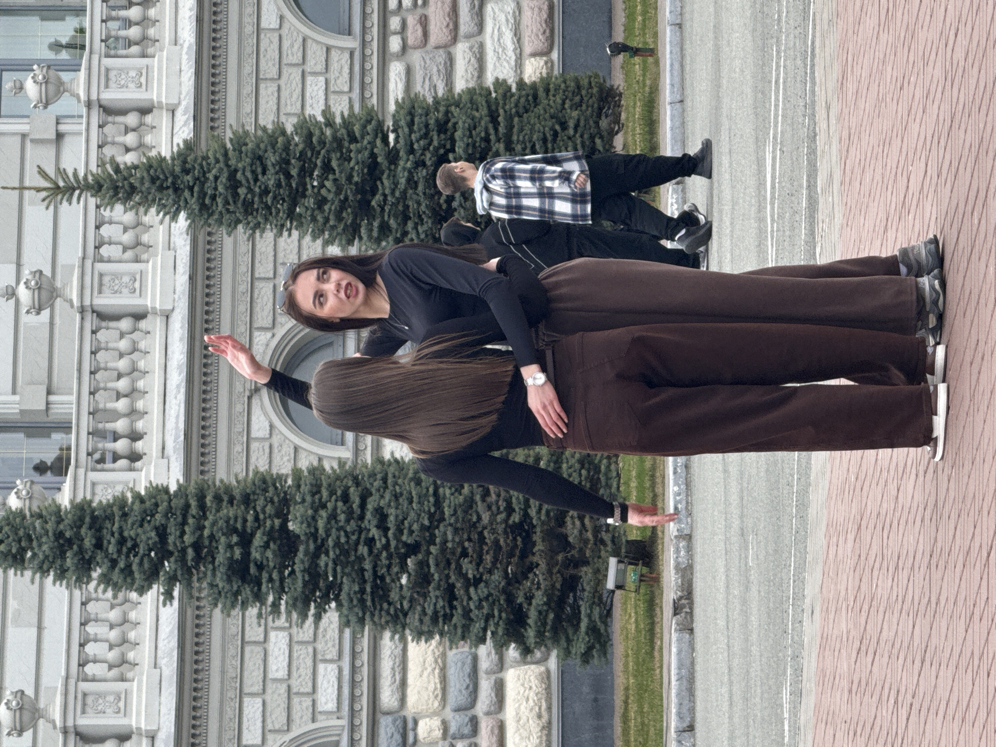 The image shows two women posing in front of a building in Kazan, Russia. The woman in the foreground is wearing a black long-sleeved shirt and brown pants. She is standing with her back to the camera and has long brown hair. The other woman is in front of her, and is wearing similar clothes. She has brown hair and is gesturing with her hand. Behind them, there is a stone building with garlands of evergreen trees hanging from the windowsills. A man in a plaid shirt can be seen walking in the background. The ground is paved with bricks and there is a street next to the building.