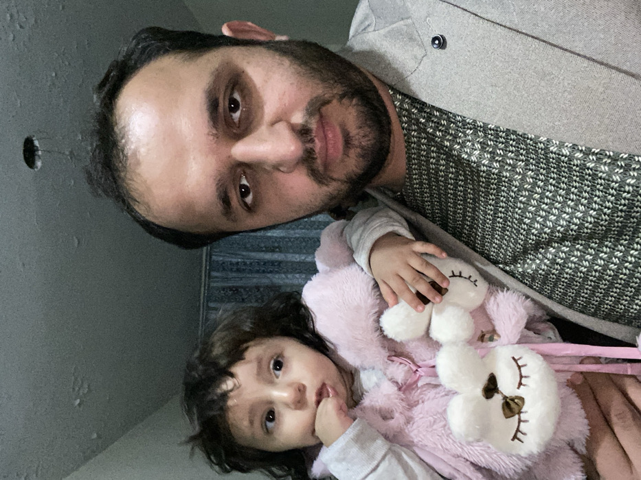 A man with dark hair and a beard poses with a young child wearing a pink fuzzy coat. The child clutches a stuffed animal resembling a bunny. The man wears a grey blazer over a patterned shirt. They are indoors with a visible wall in the background that has a hole.