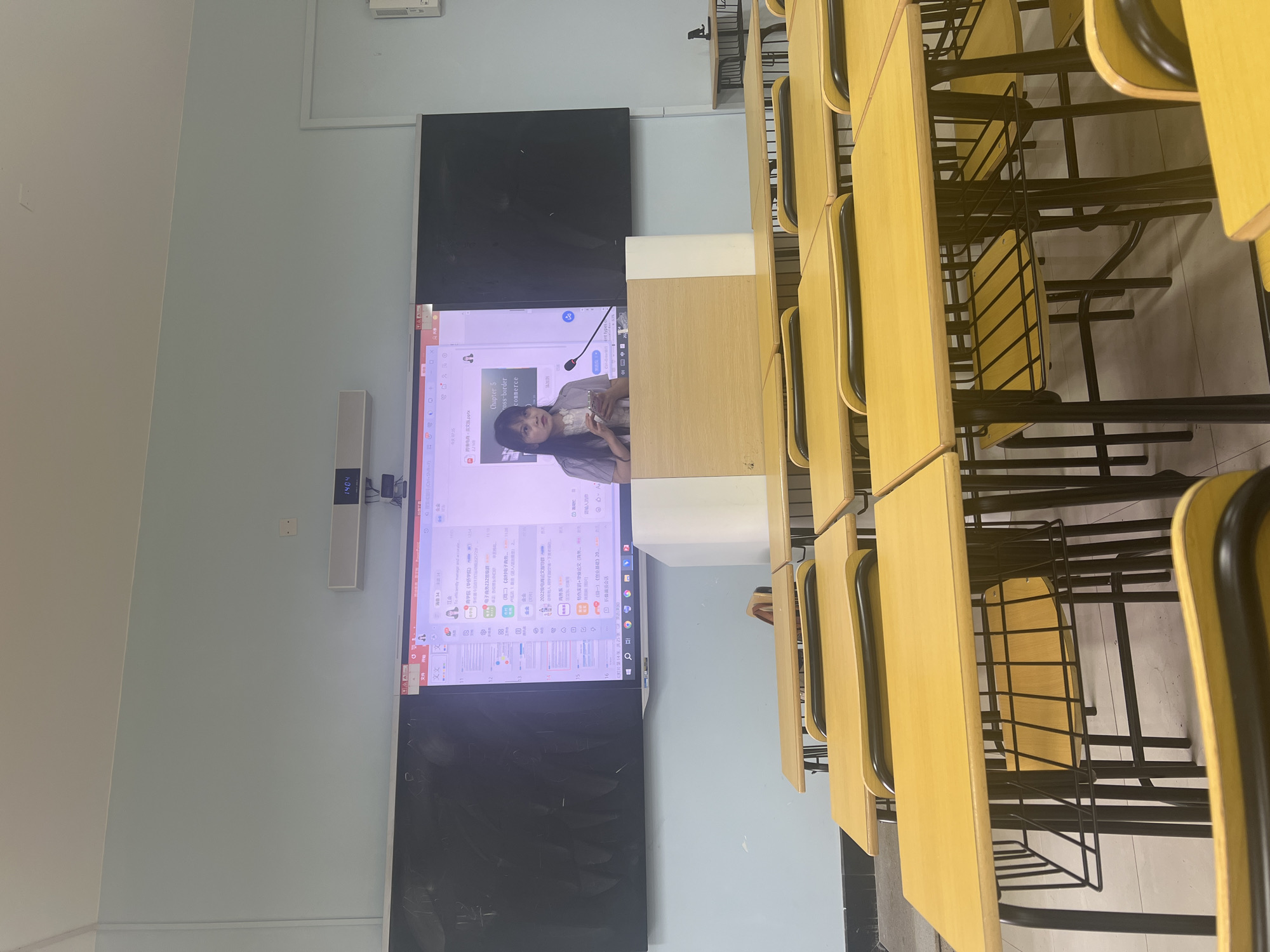 This is a photograph taken inside a classroom, likely in Liandu District, China, as evidenced by the Chinese text visible on the screen and a blackboard. The room is set up with rows of yellow-topped desks and chairs, all facing a large screen at the front of the room. A female presenter, dressed in a light-colored blouse, is standing behind a wooden podium, holding a smartphone and appearing to be in the middle of a presentation. The screen displays a presentation slide titled "Chapter 5 Cross-border commerce." The blackboard behind the screen is mostly black, with some faint writing that is illegible. The overall environment suggests an educational setting, with the presenter actively engaged with her audience, who are not visible in the frame.