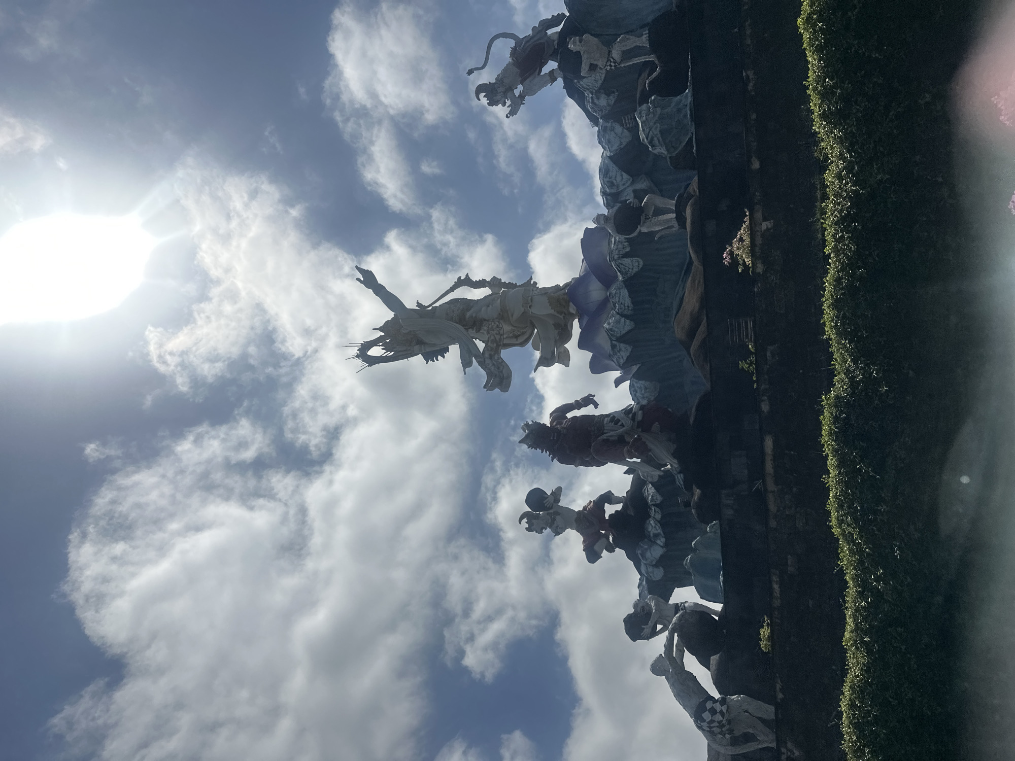 A low-angle photograph shows a tall, ornate structure against a partly cloudy sky. The structure is adorned with multiple statues of people and mythical creatures. To the right of the structure is a tall green hedge. The photograph was taken in the city of Denpasar, Indonesia.