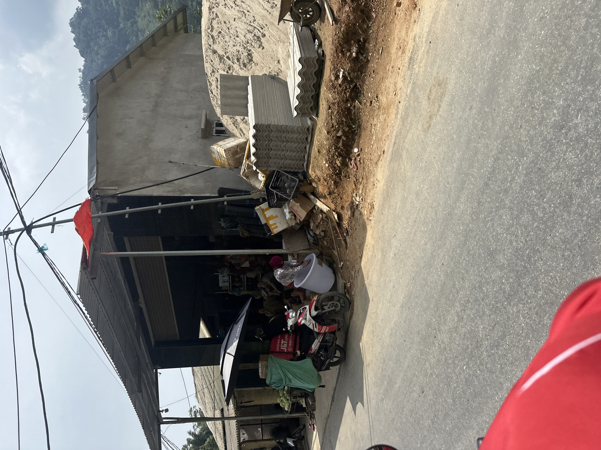 This image captures a roadside scene in Minh Sơn Commune, Vietnam, depicting a commercial area adjacent to a paved road. The main subject is a single-story concrete building with a corrugated metal roof extension, possibly serving as a shop or dwelling. To its right, a large pile of sand or dirt is visible, alongside stacks of corrugated fiber-cement roofing sheets and a wheelbarrow, suggesting construction material sales or an ongoing project.
In front of the shop, a red and white Yamaha Exciter-style scooter is parked, fitted with a red J&T Express delivery box on the back. The box prominently displays the "J&T Express" logo, along with "DỊCH VỤ PHÁT NHANH" (Express Delivery Service) and a phone number "1900 1088". Other objects near the scooter include a white plastic tub, various cardboard boxes, and a green tarp covering some goods, possibly produce like coconuts or melons. Utility poles and wires are visible in the upper left, and an orange flag hangs from the shop's roof structure.
The background features lush, tree-covered mountains under a bright, clear sky, indicating good weather, likely mid-day. The road in the foreground shows a dirt shoulder blending into the asphalt surface. The overall scene suggests a busy, functional setting common in rural or semi-urban Vietnamese areas, bustling with daily commerce and activity. The red object in the immediate foreground right is likely part of the viewer's motorcycle.