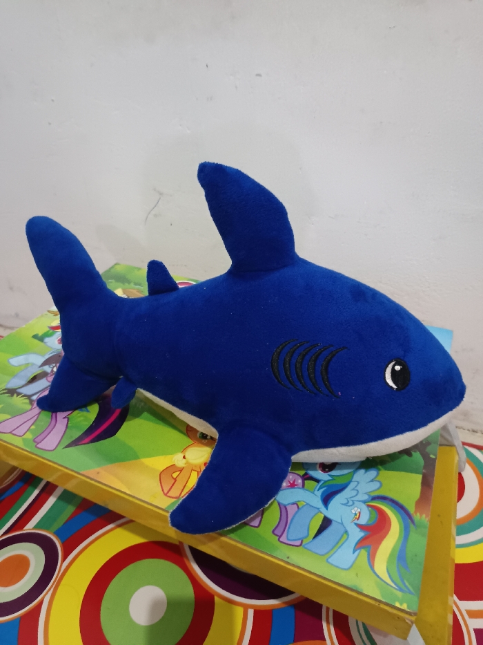 A blue plush shark toy is positioned horizontally, facing right, on a yellow-edged surface. The shark features a white underside, black stitched gill markings on its side, and black and white eyes. This yellow-edged surface is decorated with colorful illustrations of cartoon ponies, including one resembling Rainbow Dash. Beneath this surface, a multi-colored fabric with concentric circular patterns in red, orange, yellow, green, blue, purple, and white is visible. The background consists of a plain, light-colored wall with minor surface imperfections. The scene is indoors. The specific location in Gotha, Germany, cannot be determined from the visual information provided.