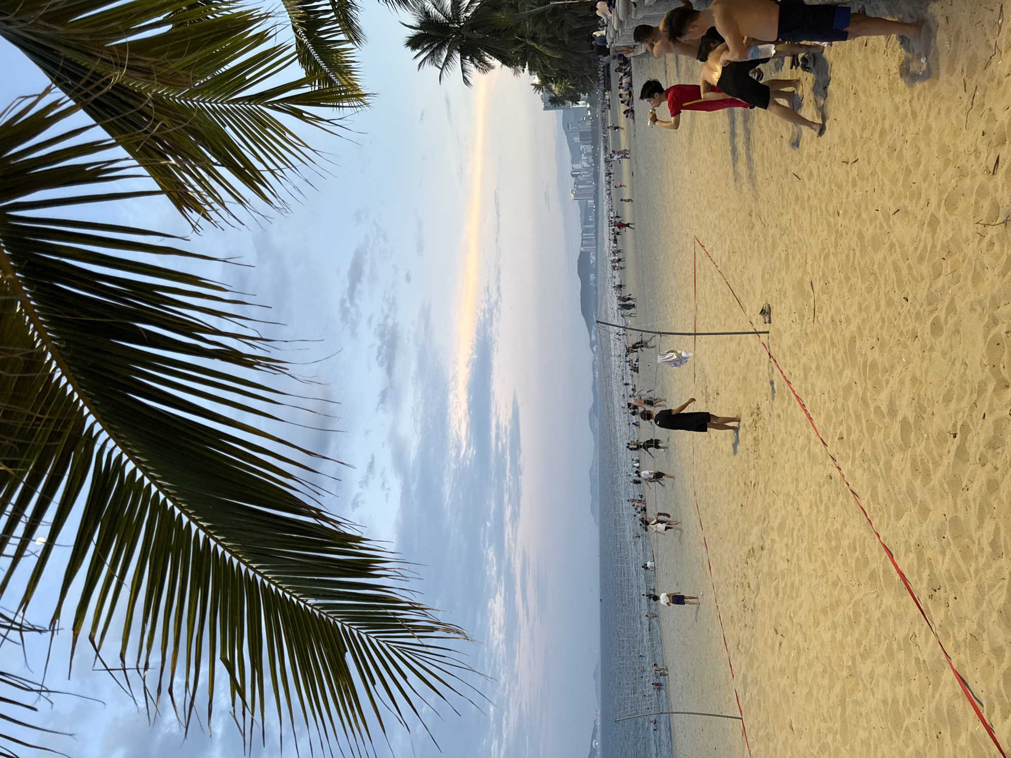 The image captures a wide view of a crowded beach in Nha Trang, Vietnam, during dusk. In the foreground, a volleyball net is set up on the sandy beach, marked with red lines. Several people are engaged in activities on the beach, including playing volleyball and standing around. Some individuals are visible in the ocean, swimming or enjoying the water. Palm trees line the edge of the beach, with their fronds extending into the frame from the top. In the background, a city skyline with tall buildings is visible, situated along a coastline, and mountains can be seen in the distance. The sky displays a soft gradient of colors, transitioning from a pale yellow and orange near the horizon to a light blue with scattered clouds. The overall scene depicts a lively beach environment in Nha Trang at the end of the day.