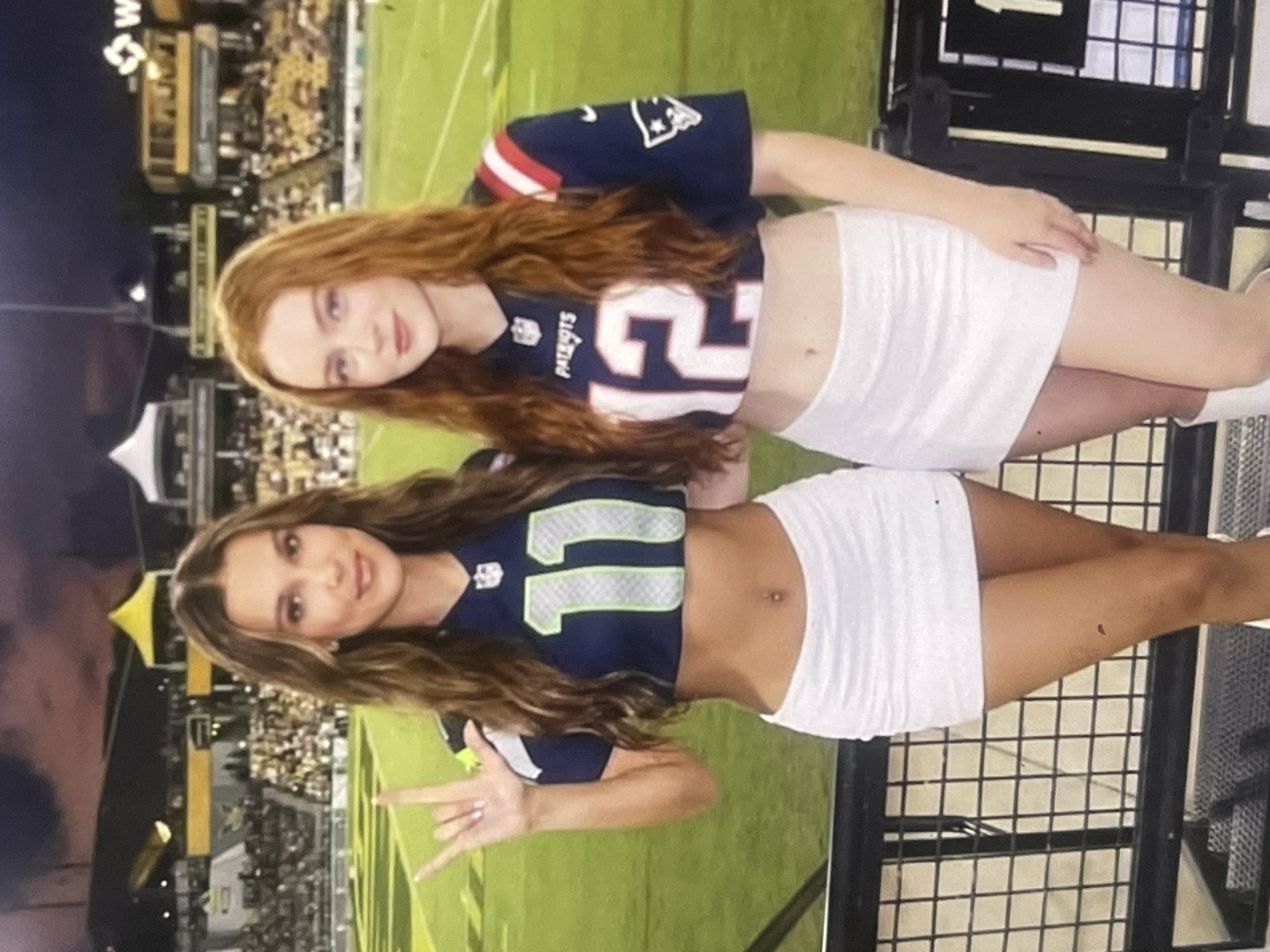 The image displays two young women posing in what appears to be an American football stadium during an evening or night event. The background shows illuminated stadium structures and stands filled with spectators, with a green field visible.
The woman on the top has long, wavy red hair and is wearing a dark blue crop top jersey with "Patriots" and the number "12" printed on it in white, along with red and white sleeve accents. She is also wearing a white mini skirt.
The woman on the bottom has long, wavy brown hair and is wearing a dark blue crop top jersey with the number "11" in light green, outlined in white. She is also wearing a white mini skirt and is making a "V" or peace sign with her left hand. Both women appear to have neutral or pleasant expressions.
The visible text on the jerseys includes "Patriots," "12," and "11." A partial letter, possibly "W" or "M," is visible on a sign in the far background.
The attire, particularly the NFL-style jerseys, and the stadium setting are characteristic of an American football game. While the provided location context is Poltava, Ukraine, the visual evidence in the image strongly suggests an American football stadium and culture, which is not typically found in that region.