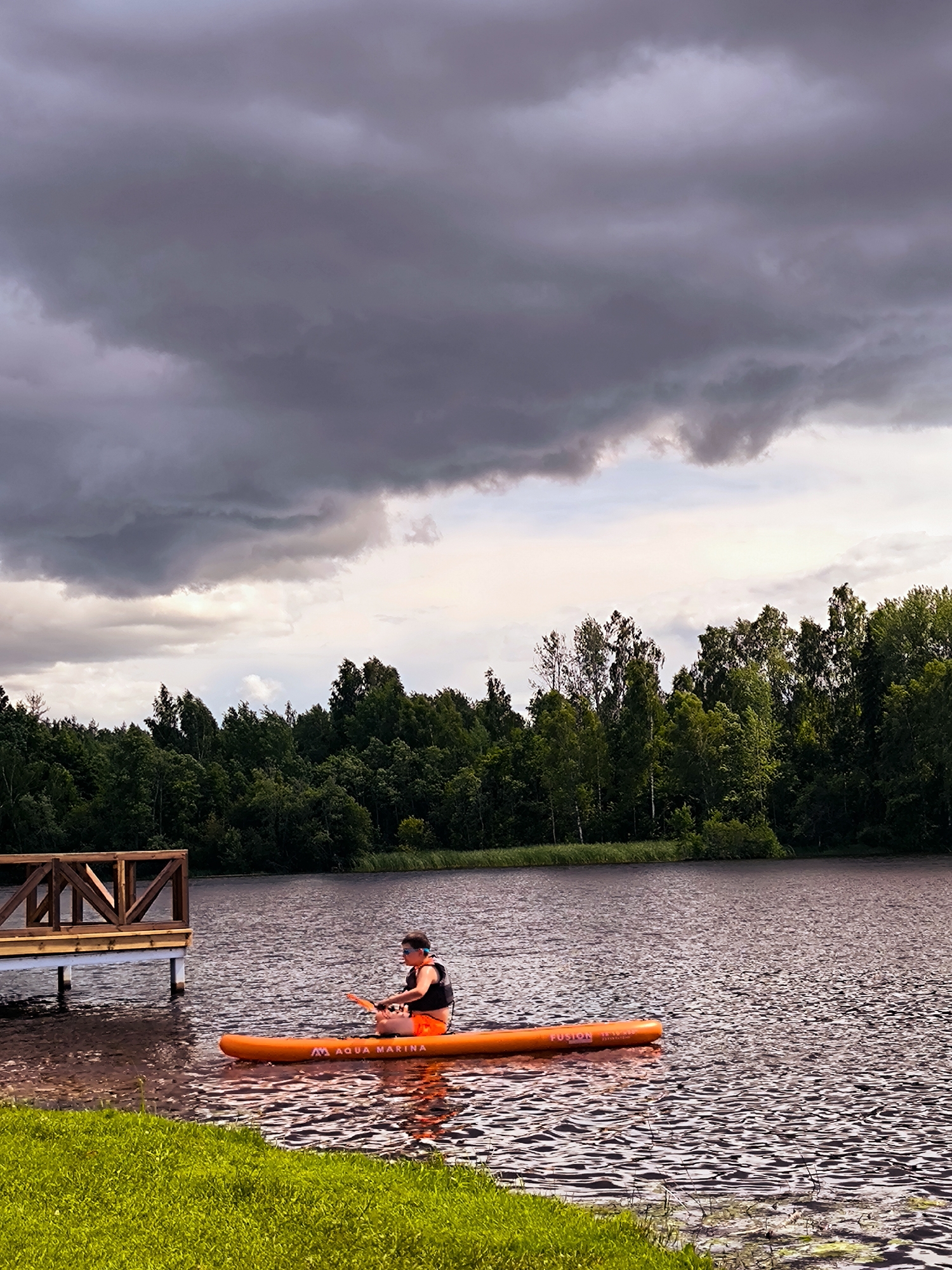An individual wearing an orange life vest and black shorts is paddling an orange stand-up paddleboard on a body of water. The paddleboard is labeled "AQUA MARINA FUSION". The person is positioned centrally on the board, facing towards the left of the frame, with a paddle in hand. In the background, a dense line of green trees forms the shoreline. To the left, a wooden dock with a railing extends into the water. The sky is overcast with dark, dramatic clouds, suggesting an impending storm. The foreground features a strip of vibrant green grass. The overall scene depicts an outdoor recreational activity on a lake or river with moody weather conditions. This scene is consistent with the natural landscapes found around Vecumnieki, Latvia, known for its numerous lakes and forests.