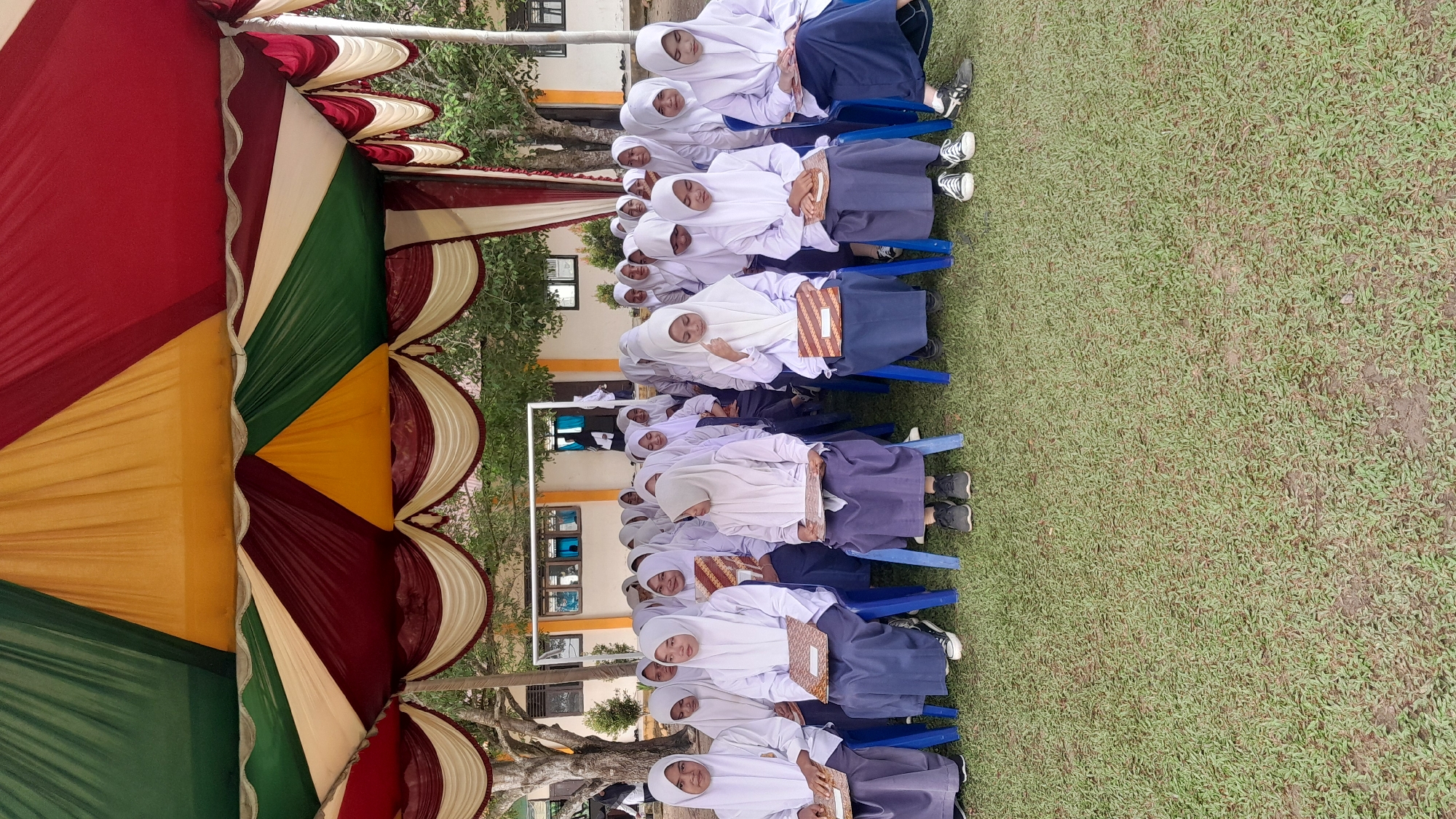 The image depicts a group of female students sitting on chairs outdoors, possibly at a school event. They are all wearing white headscarves and blue skirts, which may be part of a school uniform. The students are sitting in rows on blue plastic chairs on a grassy area. Some students are holding books or notebooks.
A colorful tent or canopy with red, green, yellow, and white fabric is visible on the left side of the image. In the background, there are buildings with windows and trees, suggesting an outdoor location possibly at Blangriek, Indonesia. The students appear to be attentive or waiting for an event to begin.