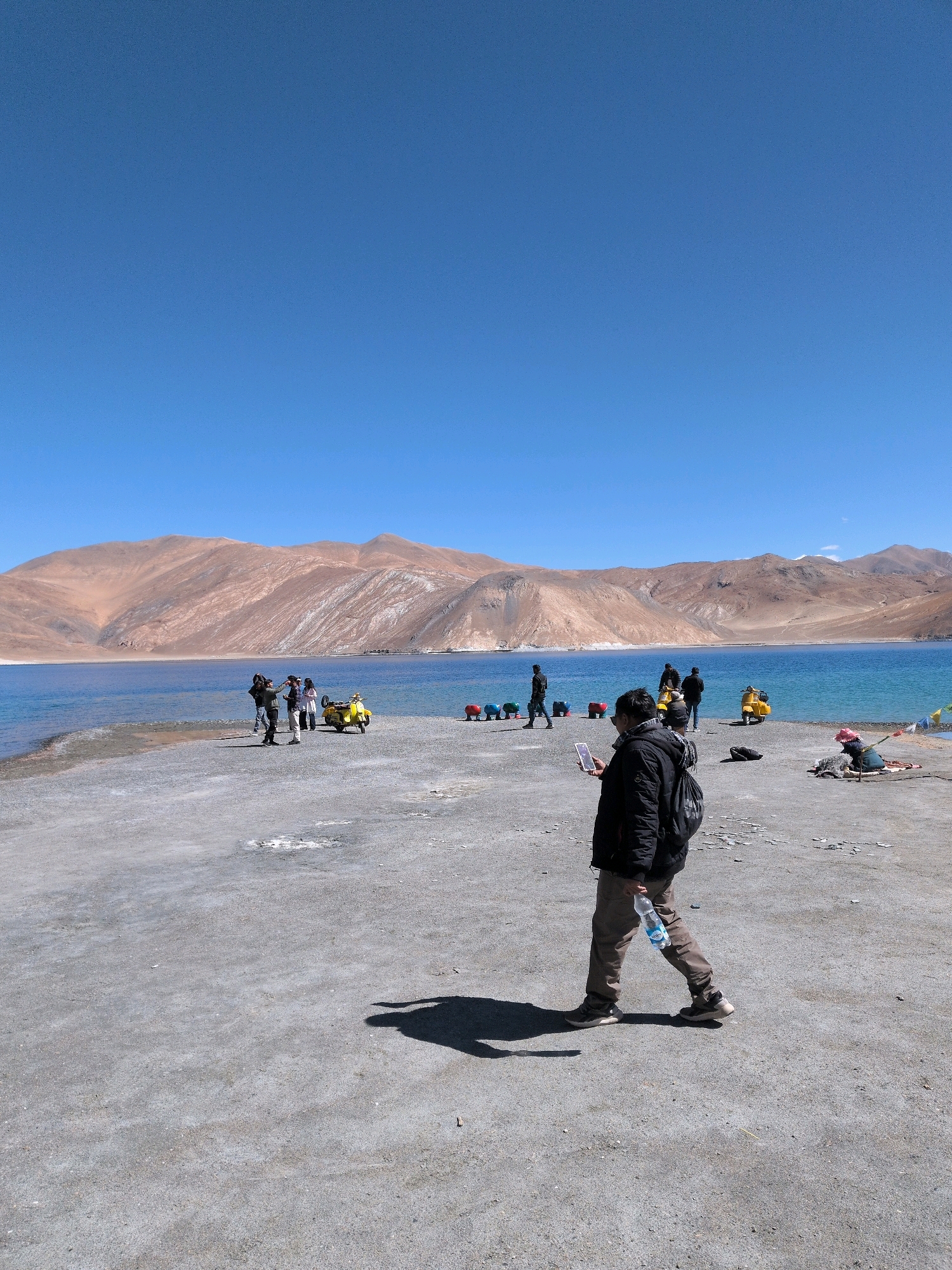 The image depicts a clear, sunny day at Pangong Tso lake, located near Leh, India. The foreground features a man walking away from the camera, holding a water bottle. He is wearing a black jacket, brown pants, and athletic shoes. His shadow is cast on the grey, pebbly ground. In the middle ground, a group of tourists are gathered near a bright yellow scooter, with several other individuals and colorful, cylindrical objects visible along the shoreline. The background is dominated by the vast, blue expanse of the lake, with imposing, arid mountains rising behind it. The sky is a vivid, cloudless blue.
