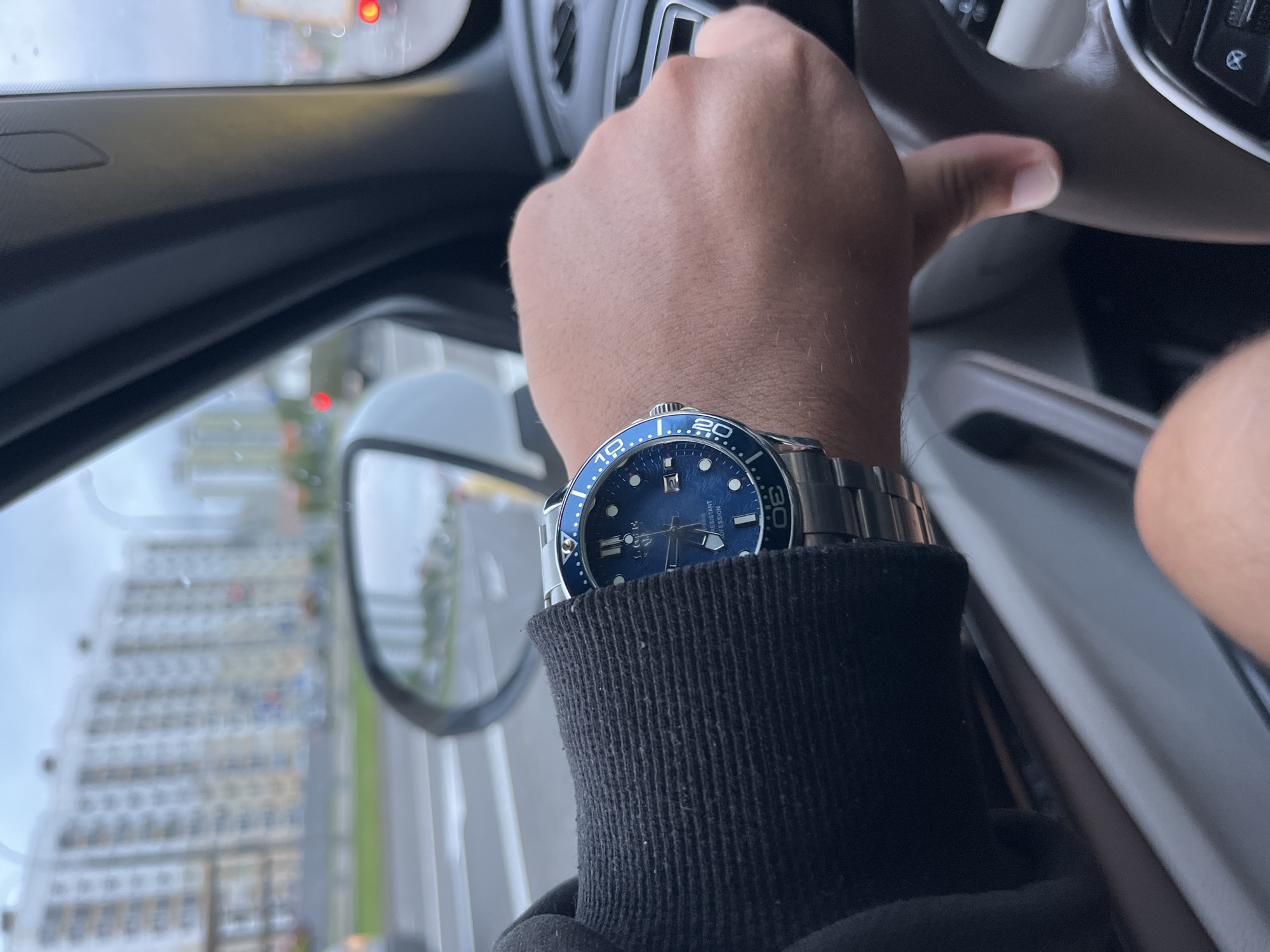 The image shows a person's arm with a watch on their left wrist, resting on the steering wheel of a car. The watch has a blue face and a metal band. The person is wearing a black long-sleeved shirt. In the background, through the car window, there are buildings and a street scene, suggesting an urban environment. The sky is overcast. The reflection in the side mirror shows more buildings and a street. The scene appears to be in the city of Minsk, Belarus, given the context provided.