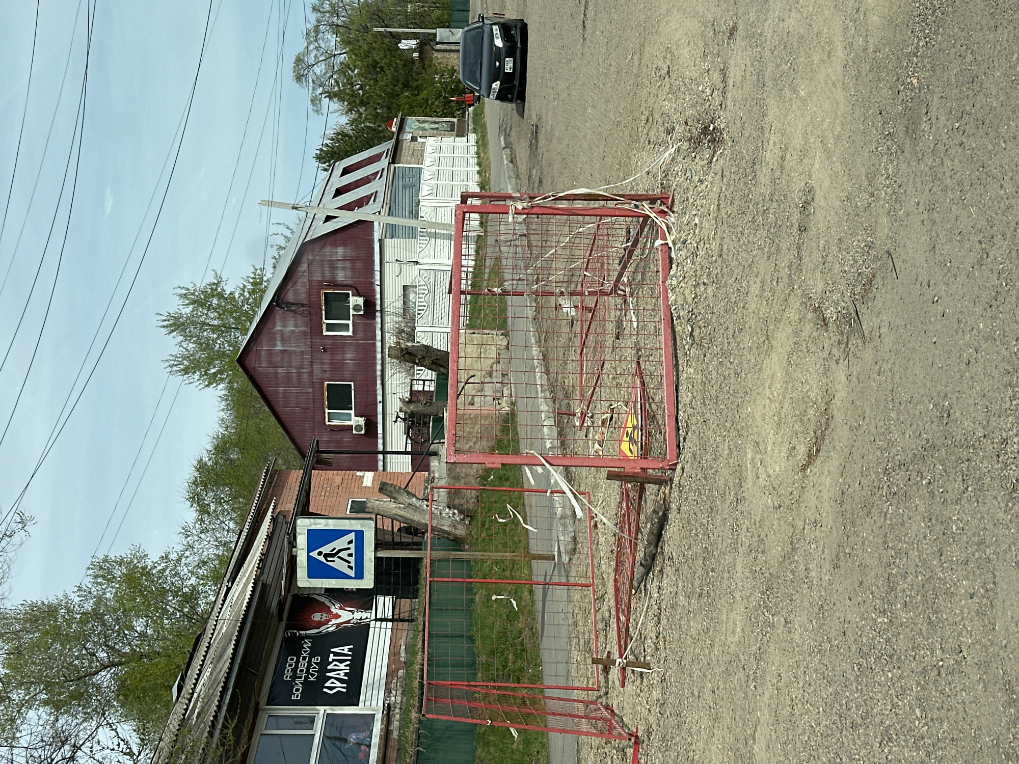 The image shows a street scene in Raychikhinsk, Russia. A red metal barrier sits in the foreground on a gravel road, secured with what appears to be white tape. Behind the barrier is a building, on which a sign that includes the words "APOO" and "SPARTA" can be seen in the lower left corner, as well as a pedestrian crossing sign. Further down the road are two more buildings, including one with a steeply pitched roof with red and white panels. In the background, a black car is parked. Overhead are power lines.