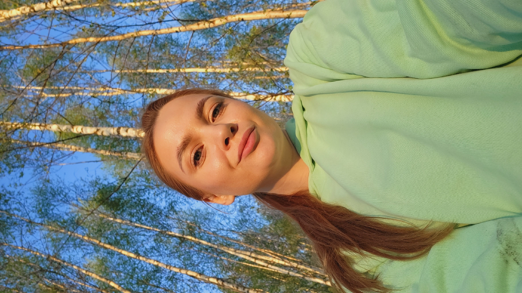 A woman with fair skin and reddish-brown hair is looking at the camera. She is wearing a light green hooded sweatshirt. The background features a stand of birch trees with white bark and green leaves against a blue sky. The picture appears to be taken outdoors, possibly in Зипуново, Russia.