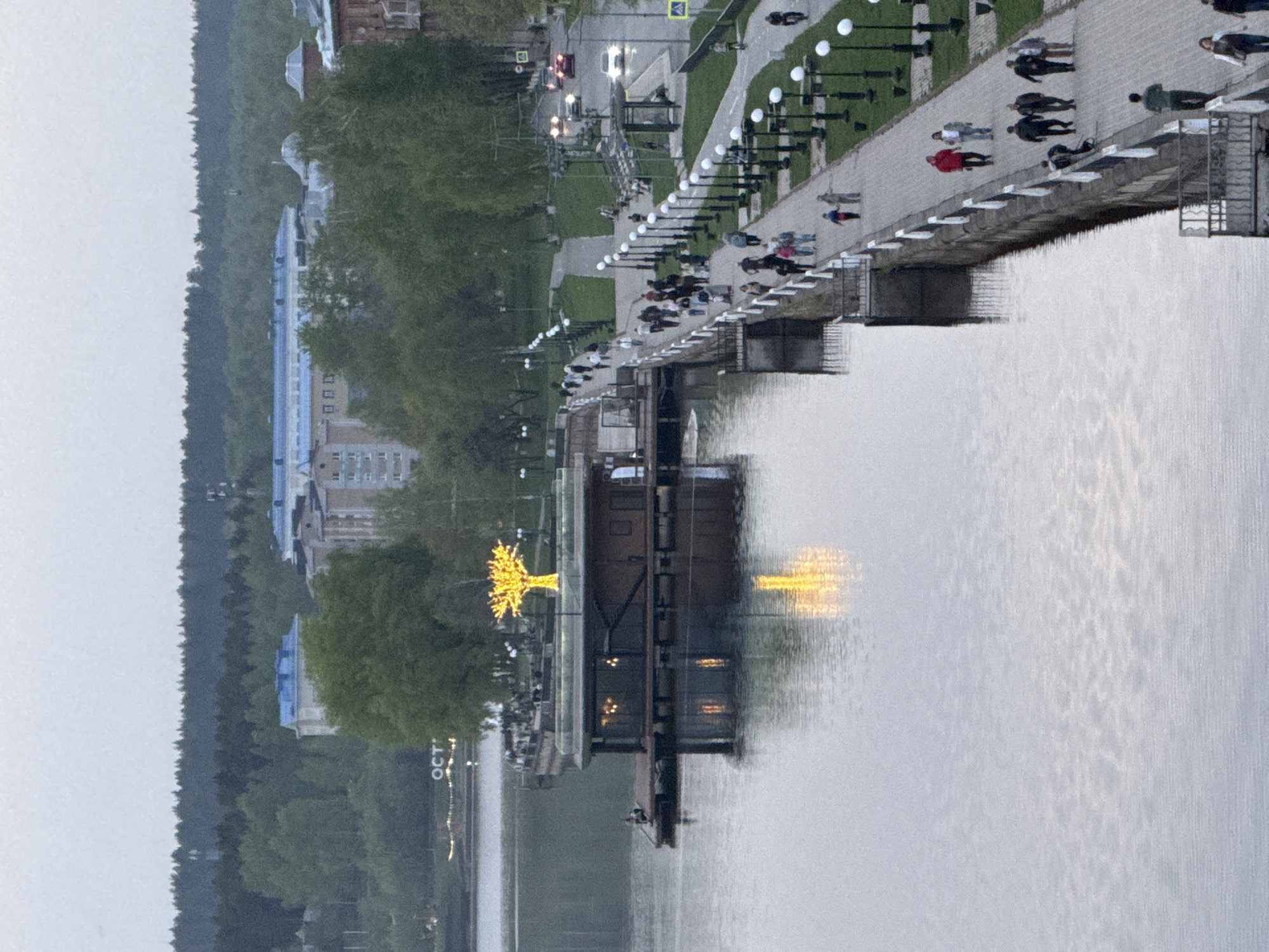 The image depicts a body of water, likely a river or lake, with a bridge spanning it. People are walking along the bridge. Adjacent to the bridge is a walkway with lampposts. The river reflects the bridge, the lampposts, and some lights from the shore. On the shoreline, there are trees and buildings. A golden tree sculpture is visible. In the background, a forest line extends along the edge of the city. The location is Votkinsk, Russia.