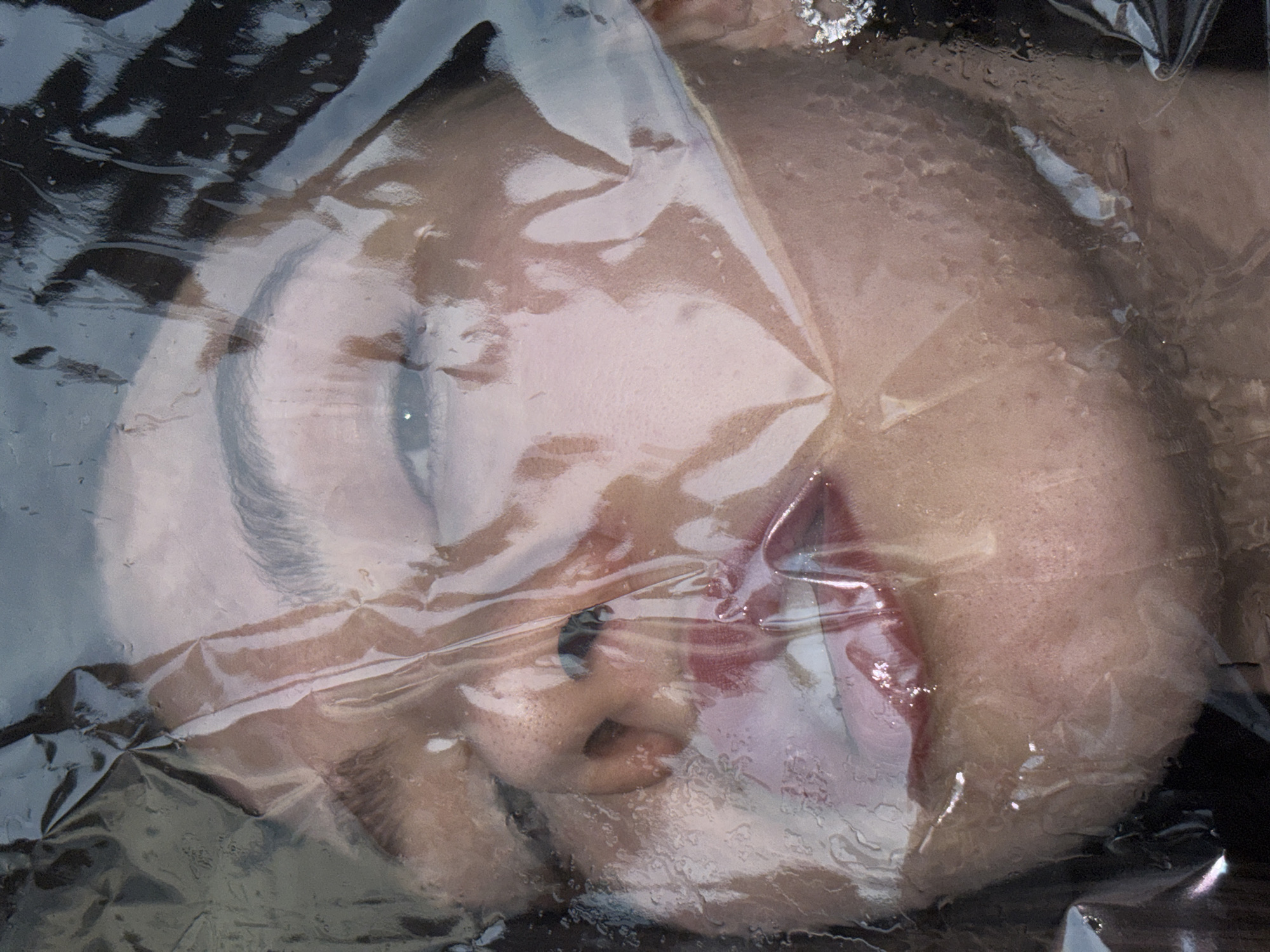 A close-up, slightly angled shot depicts a person's face covered in clear plastic wrap. The face, which is slightly distorted by the plastic, shows the eye, nose, and mouth. The plastic wrap is creased and appears damp. The background is dark and indistinct. The image does not contain sufficient detail to determine its location in the city of Perm, Russia.