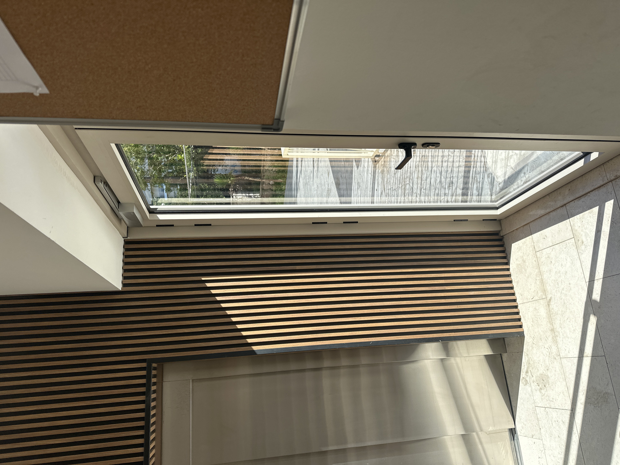 The image shows an interior view looking out through a window. The interior features a wall with horizontal wooden slats, a light-colored wall, and a silver door or panel. The window offers a view of an exterior space with a wooden deck and trees. The outdoor area appears to be sunny. The location is unspecified; the reference to Budva, Montenegro is not determinable from the visual information. There are no people visible. The environment appears to be contemporary and well-maintained. The window is open.