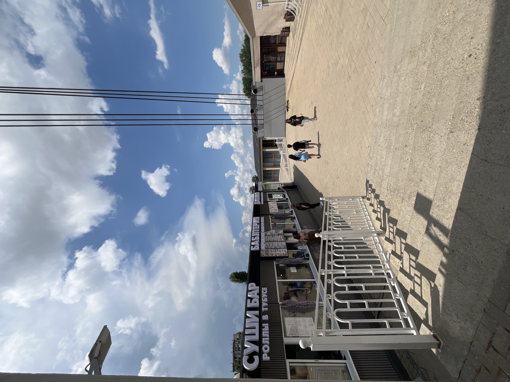 The image depicts a sun-drenched outdoor scene in Novaya Adygeya, Russia, likely near a waterfront. In the foreground, a white metal railing with an ornate design lines a set of concrete stairs casting long shadows. To the left, a building with large windows and illuminated signage identifies a "SUSHI BAR" and "ROLLS IN TUBUS." Several people are visible behind the counter inside the establishment, and some patrons are standing outside. Adjacent to the sushi bar, another establishment with signage that includes "BABEL" is visible.
Further into the scene, a paved plaza stretches out, populated by a few individuals walking. Three vertical dark lines, possibly part of a structure or overhead cables, dominate the upper central portion of the image against a backdrop of a bright blue sky with fluffy white clouds. In the distance, more buildings and trees form the horizon. The overall atmosphere is casual and suggests a public recreational area or promenade.