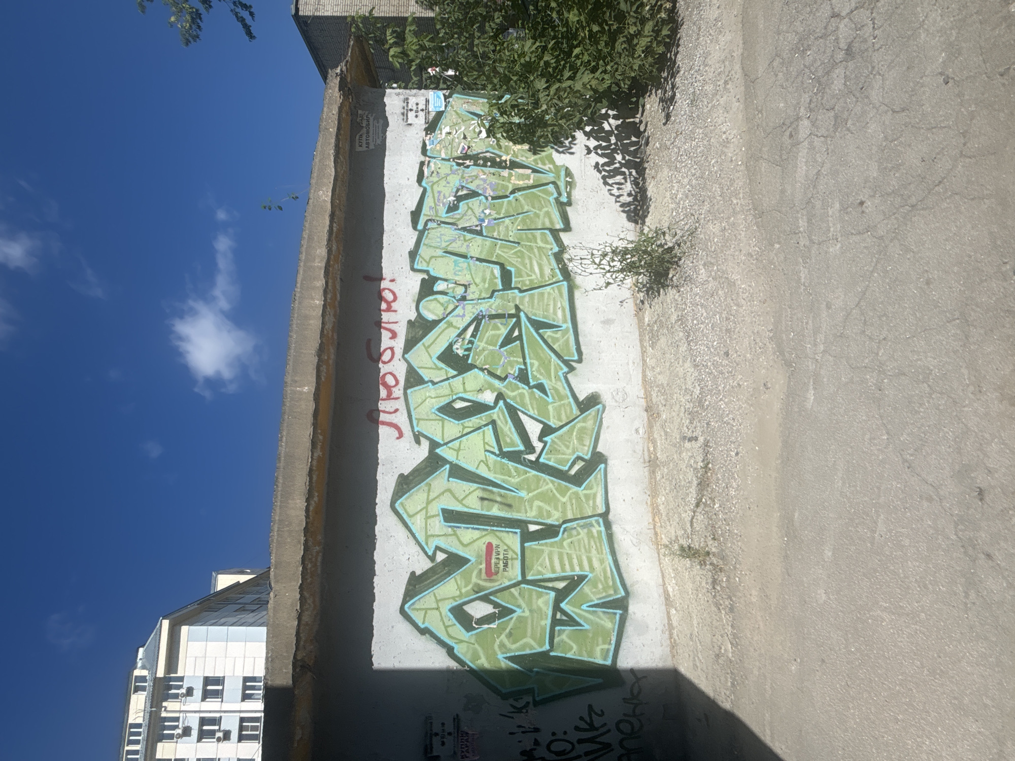 The image shows a wall with graffiti in Novosibirsk, Russia. The graffiti is in light green with a blue outline and depicts stylized letters. Above the graffiti, there is red text that reads "Люблю!". To the right of the graffiti, there is a small sign with text that is difficult to read, but appears to say "КУПЛЯ", "ВАШ", and "АВТОМОБИЛЬ". A patch of green foliage is visible to the right of the wall, and a modern building is in the background to the left. The ground in front of the wall is paved with asphalt and gravel. The sky is blue with scattered white clouds.