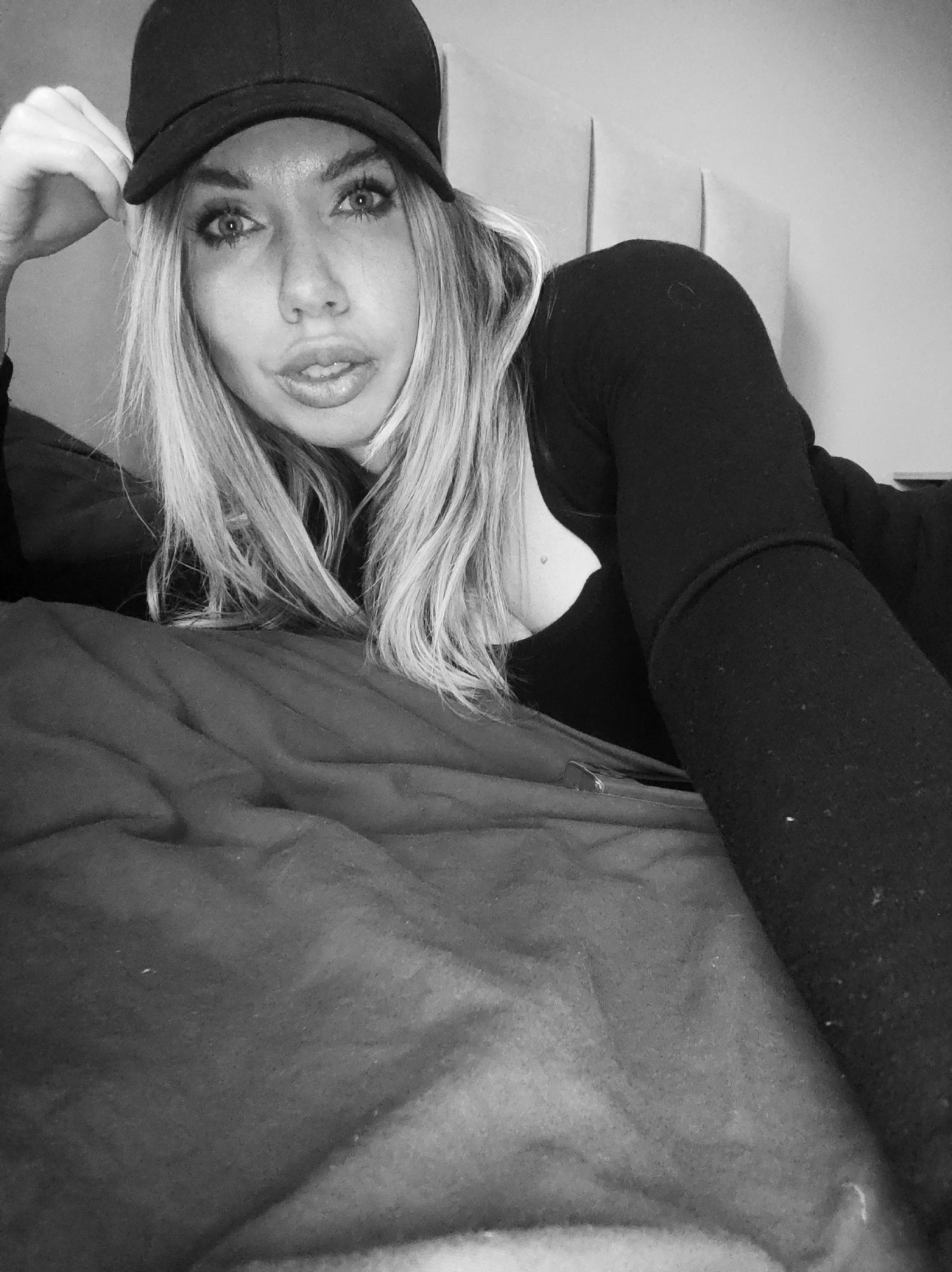 The monochromatic image features a young woman with long, light-colored hair, wearing a dark baseball cap and a dark, long-sleeved top. She is lying down, resting on her arm, on what appears to be a bed or sofa covered with dark, rumpled fabric. Behind her, light-colored, possibly padded wall paneling or a headboard is visible.
The woman is looking directly at the camera with a direct and somewhat intense gaze, her eyes wide open and her lips slightly parted. Her left hand is raised, lightly touching the brim of her cap. A small, light-colored mark is visible near her left collarbone. The lighting is soft and even, indicating an indoor setting. No specific time of day or weather conditions can be discerned, and there are no visible location cues within the image itself beyond it being an indoor environment. There is no visible text. The activity captured is a posed portrait or selfie. This scene is located in Votkinsk, Russia.