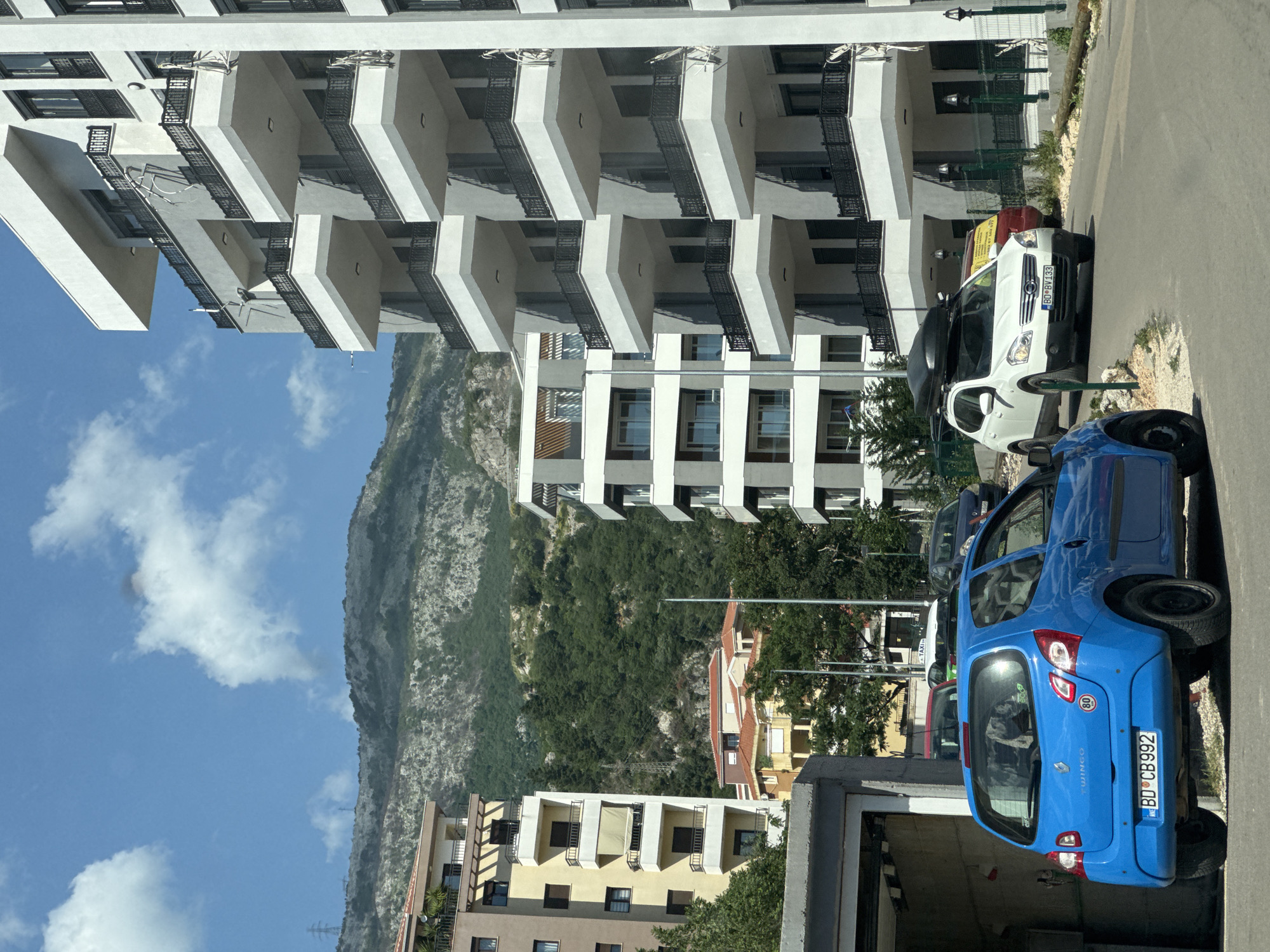 The image shows a street scene in Budva, Montenegro. On the right side of the frame are a blue Renault Twingo (license plate BD-CB992) and a white Opel Combo (license plate BD-BY133) parked in front of a building. Behind the cars are a multistory building with multiple balconies. In the background are the mountains of Budva. The sky is blue with scattered clouds. Other buildings are visible in the midground.