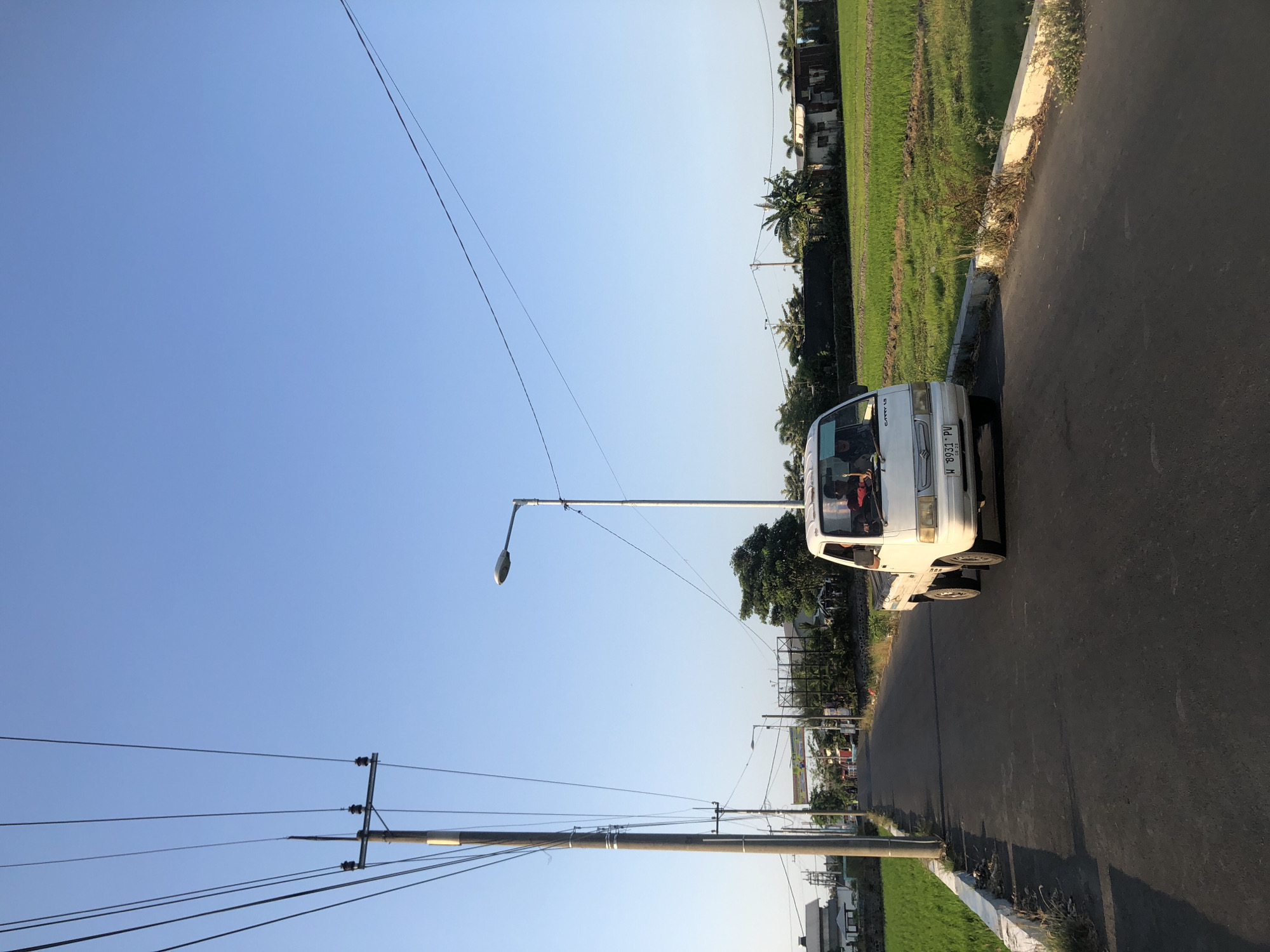 A white Suzuki Carry pickup truck with a license plate displaying "W 8931 PV" is driving down a paved road in Kalitengah, Indonesia. The truck has two visible occupants. The road is lined with a concrete barrier on the left, separating it from a vibrant green rice paddy field. On the right side of the road, beyond the truck, are more rice paddies and various buildings, including houses and what appears to be a structure with corrugated metal roofing. Overhead, a clear blue sky is crisscrossed with numerous power lines, supported by utility poles. One prominent light post stands to the right of the road. The scene depicts a rural or semi-rural setting within Kalitengah.