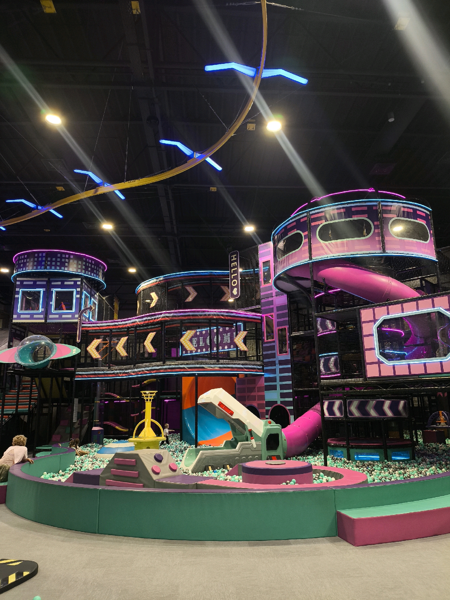 The image depicts an indoor playground with a futuristic, neon-lit theme. The structure is multi-level, featuring tubes, slides, and climbing areas. Dominant colors include purple, pink, teal, and blue neon accents. A ball pit filled with light green and grey balls is situated at the base of the play structure. Several children are visible, with one child sitting on the edge of the ball pit in the foreground, and others playing further inside. The environment appears to be a large, open indoor space with a dark ceiling and overhead lighting. The overall impression is one of a vibrant and engaging play area. There is no discernible information in the image to associate it with the city of Орловка, Russia.