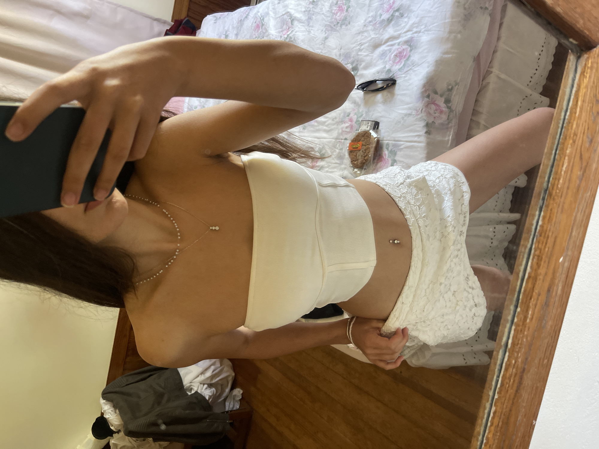 A young woman is captured in a mirror selfie in an indoor environment. She is wearing a white strapless top and a white lace skirt. She is accessorized with a delicate pearl necklace and a belly button piercing. Her left hand is holding the edge of her skirt, lifting it slightly. On the bed behind her, there is a cookie, a pair of sunglasses, and a phone. To the left of the frame, a jacket is draped over a piece of furniture. The overall impression is of a casual, personal moment in what appears to be a bedroom. The image is consistent with a scene that could be found in Oak Bluffs, Massachusetts, United States of America, a popular vacation destination known for its charming cottages and relaxed atmosphere.