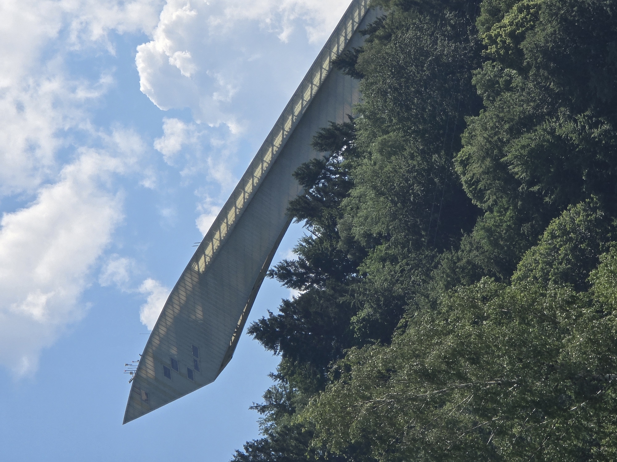 The media file shows a ski jump in Garmisch-Partenkirchen, Germany. The ski jump is a large, modern structure with a metallic finish, angled against a bright blue sky with scattered white clouds. It is surrounded by lush green trees, suggesting a summer or early autumn setting. The perspective is from below, looking up at the imposing structure.
The ski jump appears to be empty, with no athletes or spectators visible. This implies that there is no current event taking place, such as a competition or training session. The lighting suggests it is daytime, likely midday or early afternoon, with the sun casting a bright, even light. The weather appears to be clear and pleasant.
Notable details include the intricate scaffolding or support structure visible along the edge of the ski jump's takeoff ramp, and several small windows or openings on the side of the structure near the bottom. The overall scene conveys a sense of scale and architectural grandeur within a natural setting. There is no visible text within the image.