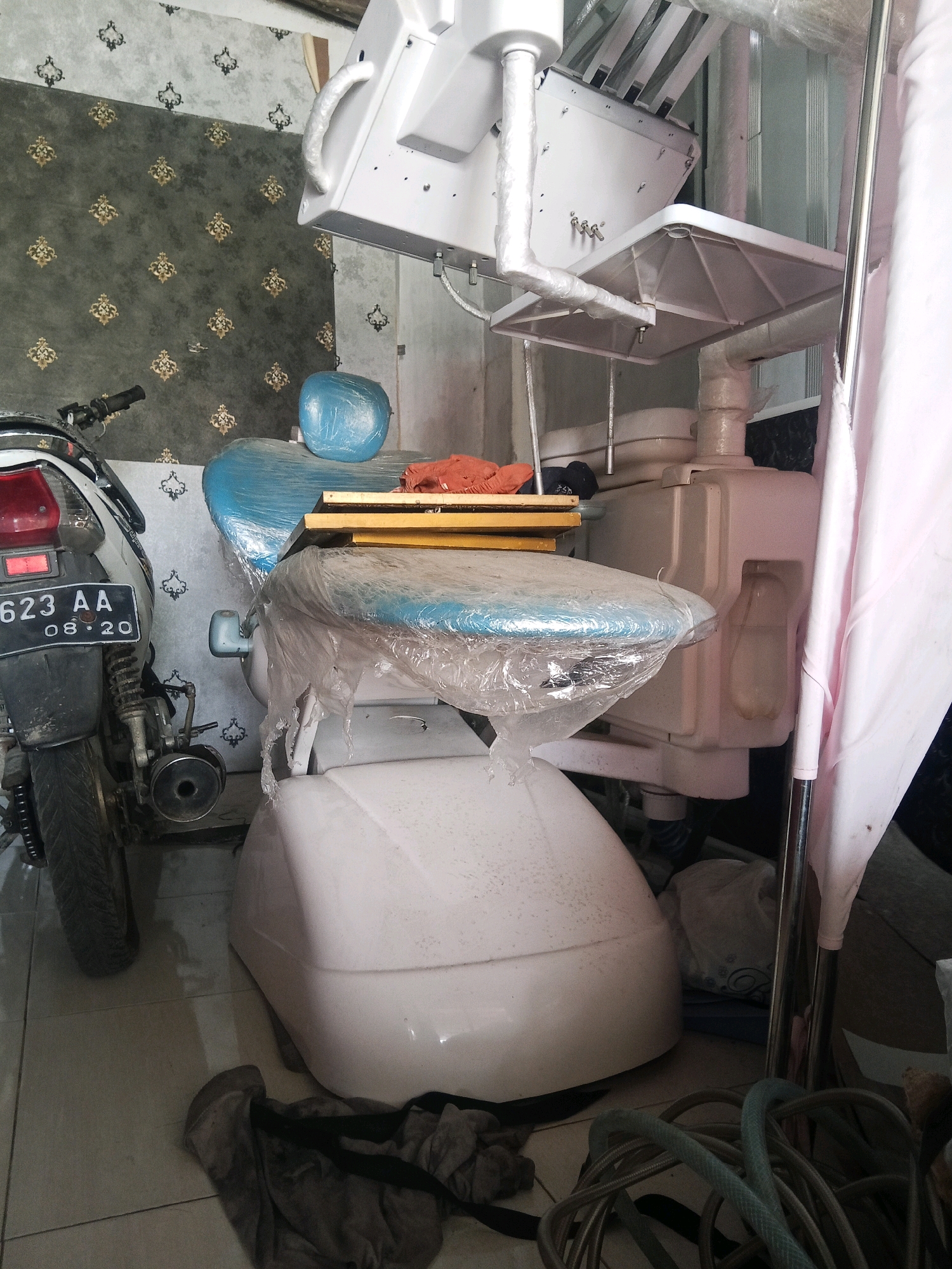 In the city of Jinang Kampung Pansur, Indonesia, a dental chair is positioned in what appears to be a storage area or a workspace. The dental chair is white and covered in plastic wrap, as are parts of the surrounding equipment. A motorcycle, with license plate "623 AA 08.20," is parked to the left of the chair, also partially covered in plastic. In the background, a patterned wallpaper is visible. Various items, including a blue headrest, orange cloth, and coiled hoses, are scattered around the dental chair and on the floor. The scene suggests an environment where dental equipment is either being stored, cleaned, or prepared for use.