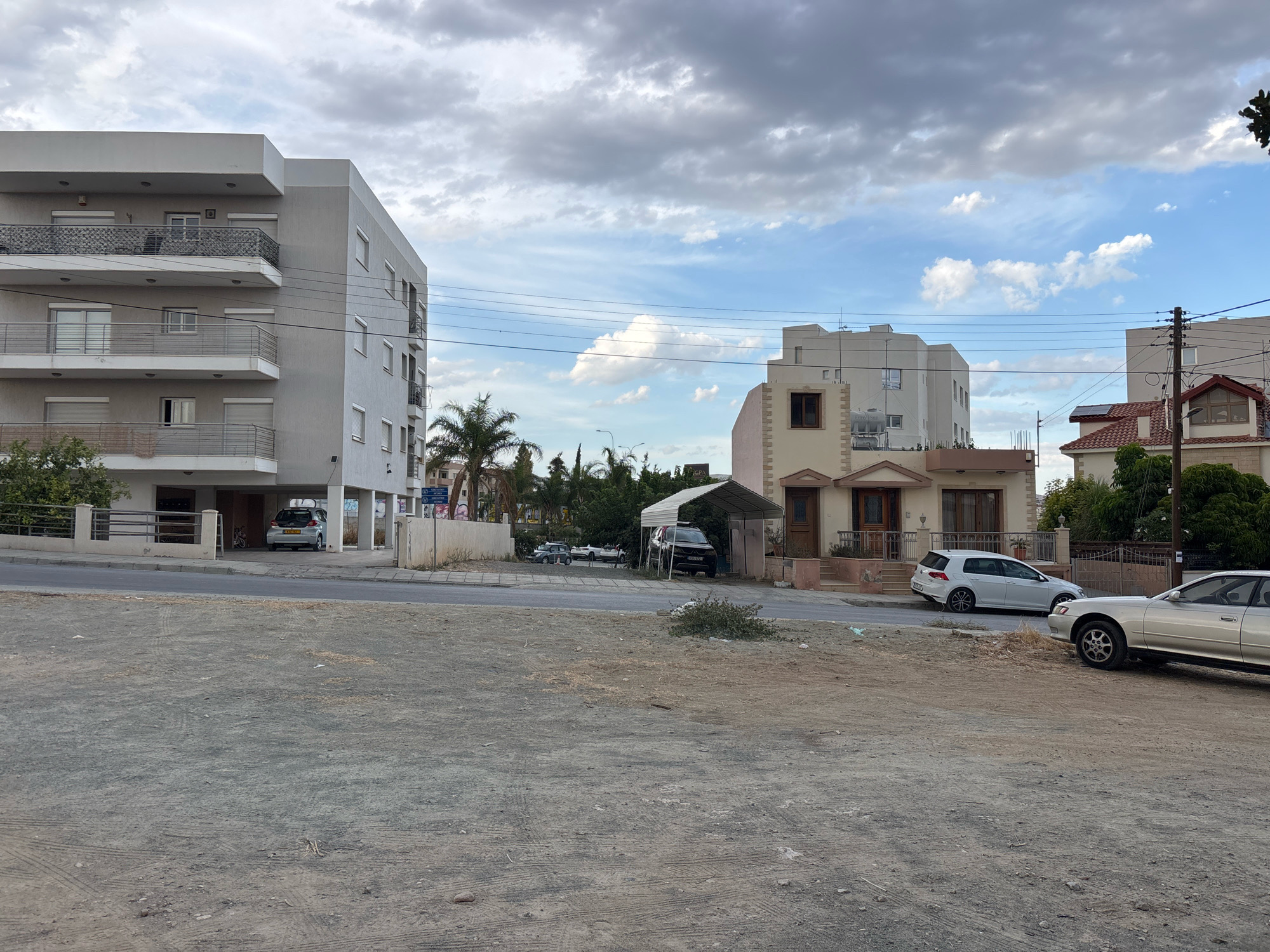 The image depicts a street scene in Mesa Geitonia, Cyprus, under a cloudy sky. On the left, a multi-story apartment building with grey siding and balconies is visible. To the right of the apartment building, several cars are parked. In the center of the image, a two-story house with a light-colored facade and a covered carport is present, with a dark SUV parked underneath. Further to the right, another house with a red tiled roof and a white sedan parked in front of it can be seen. The foreground is a dirt or gravel lot. The scene appears to be a residential street with a mix of apartment buildings and houses.