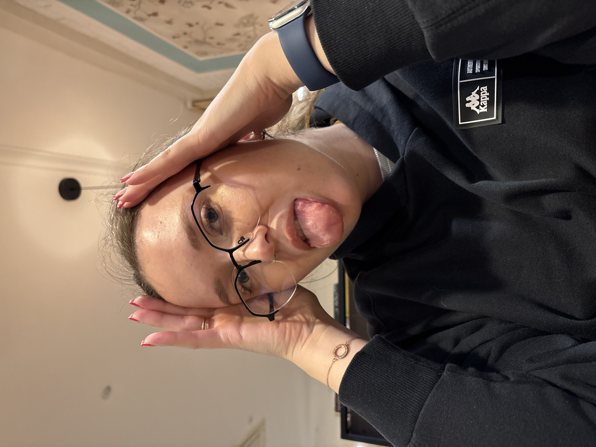 A person is positioned upside down in the image. They are wearing glasses, a black Kappa-branded sweatshirt, and a blue smartwatch. Their hands are placed on their forehead. The person is sticking their tongue out. The background includes a ceiling with a decorative pattern and a light fixture. No specific location or city is discernible from the image.