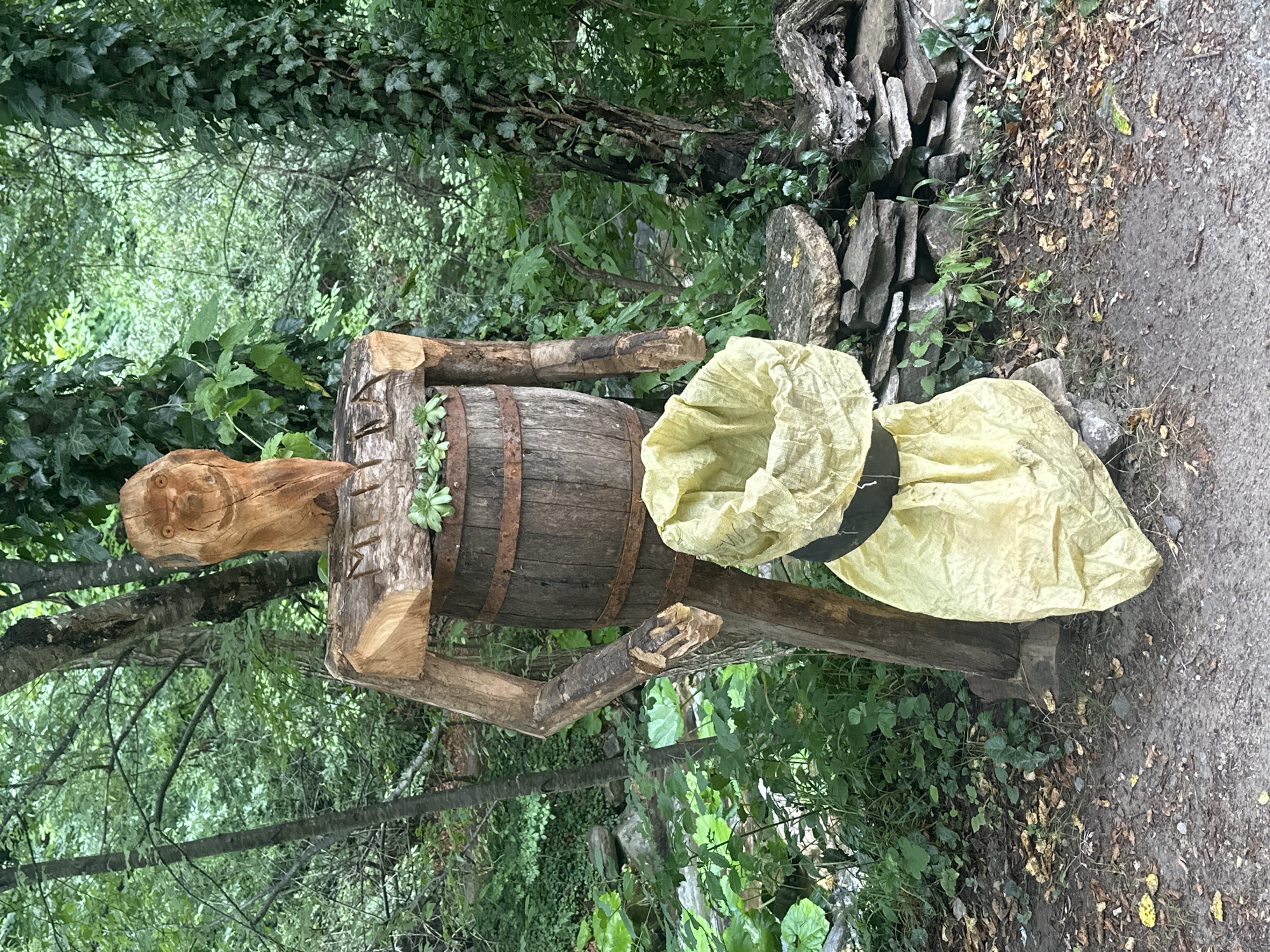 A wooden sculpture of a figure stands outdoors in a forest setting. The figure is constructed from rough-hewn wood, with a barrel serving as its torso. The head is carved from a separate piece of wood and features a smiling face with carved eyes and a wide grin. Its arms are made from branches, and it appears to be wearing a large, light-yellow bag that is cinched around its waist with a black band. The sculpture is situated on a dirt path surrounded by lush green foliage, trees, and rocks. The background shows a dense forest with ivy climbing up some of the trees. The overall impression is of a whimsical, rustic sculpture placed in a natural environment. This sculpture is located in Golyamo Belovo, Bulgaria.