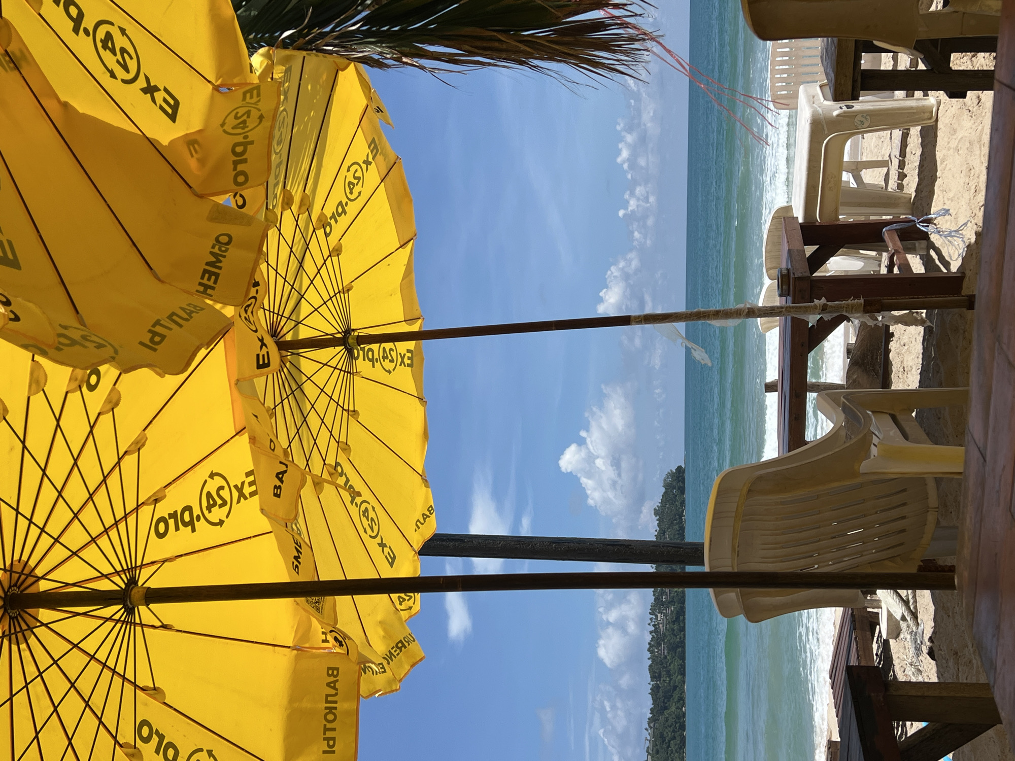 The image shows a beach scene with yellow umbrellas, chairs, and tables set up on the sand. The ocean is visible in the background with waves breaking on the shore. A lush, green coastline with buildings can be seen in the distance. The sky is blue with scattered white clouds. The scene appears to be a relaxed setting, possibly a beachfront cafe or seating area.