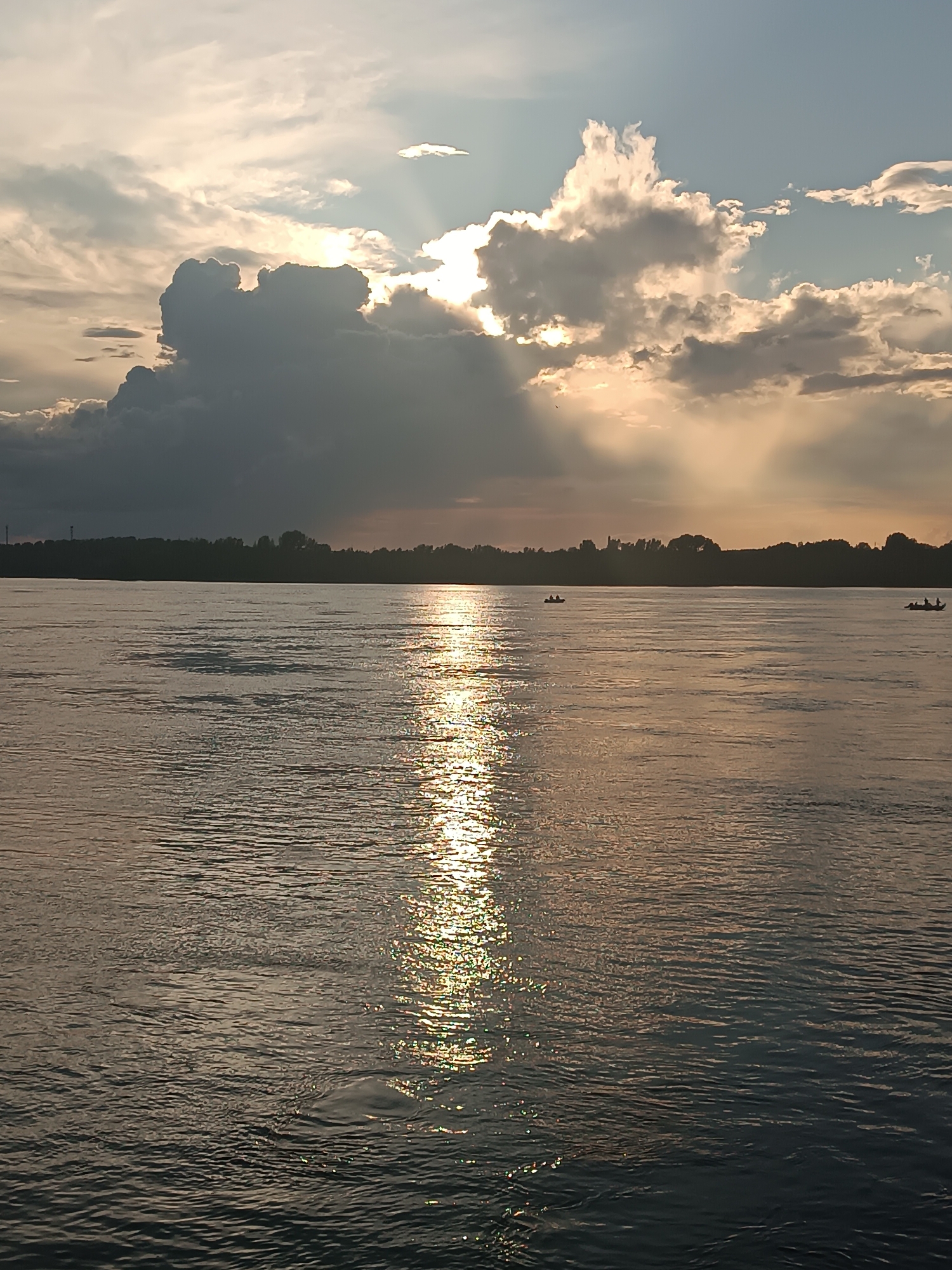 The image shows a wide, calm river with gentle ripples reflecting the golden light of the setting sun. The sun's rays pierce through dramatic, dark clouds, creating a striking contrast in the sky. A silhouetted treeline forms the distant horizon, with a few indistinct structures visible along the shore. Two small boats, each with at least one person, are on the water, adding a sense of scale and human presence to the vast natural landscape. The scene suggests a peaceful evening on the Kama River in Chaikovsky, Russia.