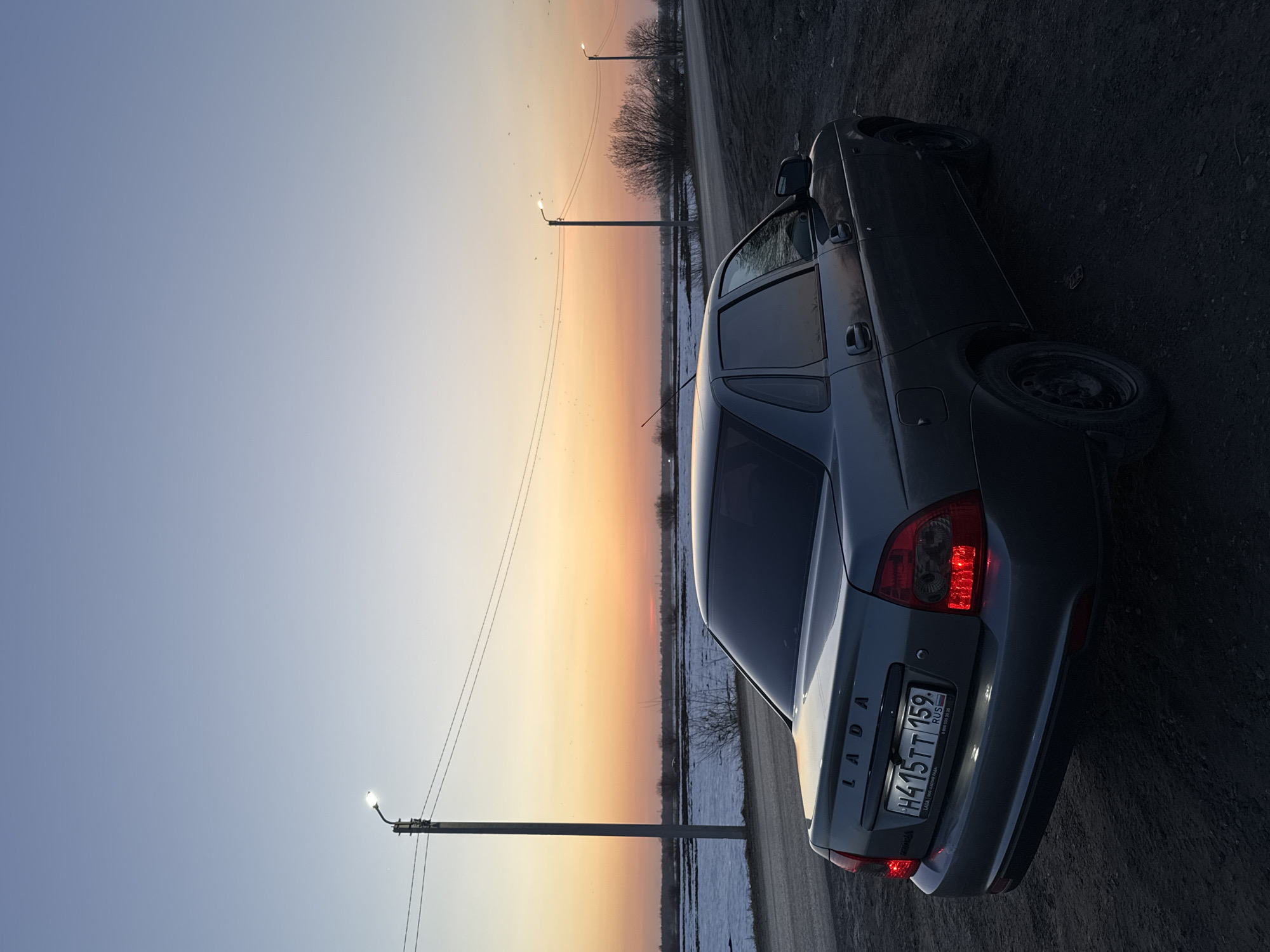 The image shows a grey Lada sedan parked on a narrow road next to a field. The rear of the car is facing the viewer. The license plate reads "H415TT 159 RUS". The background shows a sunset with colors ranging from blue to orange. There are utility poles lining the road. The picture appears to be taken in a rural or semi-rural location. While Кондратово, Russia is mentioned, its relationship to the specific image content cannot be determined with certainty.