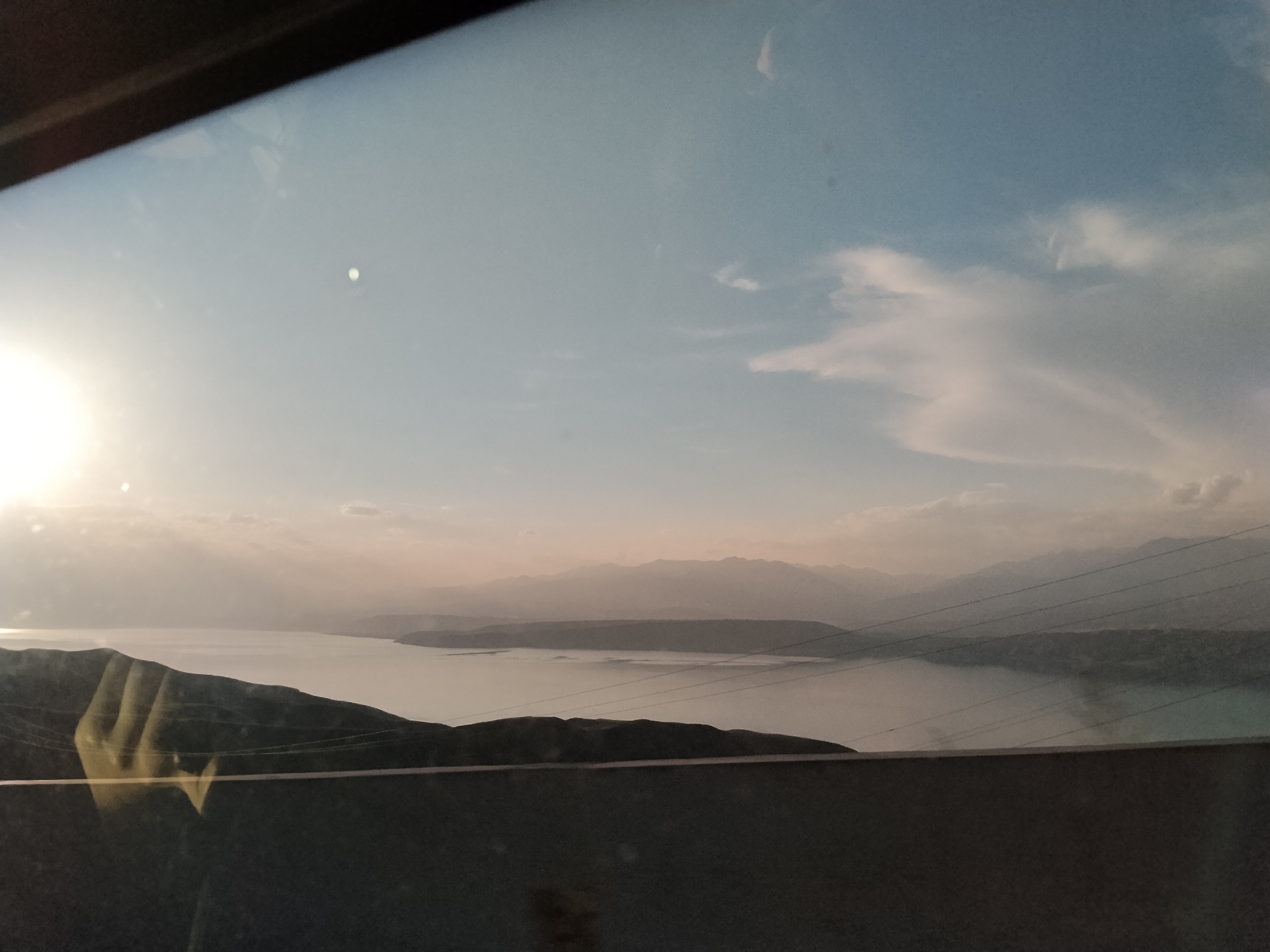 The image depicts a wide view of a large body of water surrounded by hilly terrain, viewed from an elevated perspective, possibly from a vehicle. In the foreground, a dark, horizontal structure, possibly a wall or railing, runs across the bottom of the frame. A reflection of a hand is visible on the glass through which the scene is viewed, positioned on the left side of the frame. Beyond the foreground, a body of water stretches out, with its surface appearing calm and reflecting the sky. The horizon is dominated by a series of layered mountains that fade into the distance, softened by atmospheric haze. The sky above is a gradient of pale blue to a lighter, almost white hue near the horizon, with some wispy clouds scattered across it. The sun is setting or rising, casting a warm glow and creating a bright, diffused light on the left side of the image, with its reflection visible on the water. The overall impression is one of a vast, serene landscape. The image was taken in the city of Chkalov, Kyrgyzstan.