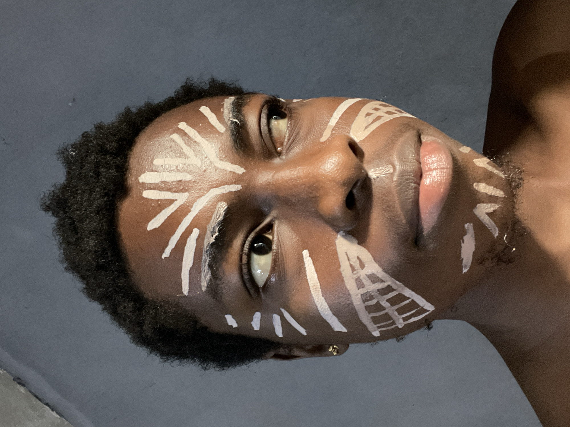 This close-up portrait, taken in Nkpor, Nigeria, features a person with dark skin and short, dark, curly hair. The individual's face and upper torso are prominently displayed, captured from a low-angle perspective that looks upwards at the subject. The person's head is tilted, and their gaze is directed upwards and slightly to the right of the frame, conveying a calm or thoughtful expression.
The most distinctive detail is the intricate white body paint adorning the face. Geometric and abstract patterns, including lines, dots, and a grid-like design on the cheek, are applied across the forehead, cheeks, and potentially extending to the neck and shoulder area. The paint varies in thickness, suggesting a hand-painted application. A single gold stud earring is visible on the left earlobe.
The background is a plain, dark grey or blue-grey surface, indicating an indoor setting or a studio environment with controlled lighting. Soft, diffused light illuminates the subject's features, creating a subtle sheen on the skin, particularly on the forehead, nose, and lips. No specific time of day or weather conditions can be determined from the image. The bare shoulder suggests attire suitable for showcasing body art. The portrait appears to capture a moment of cultural expression, performance, or artistic display. There is no visible text in the image.