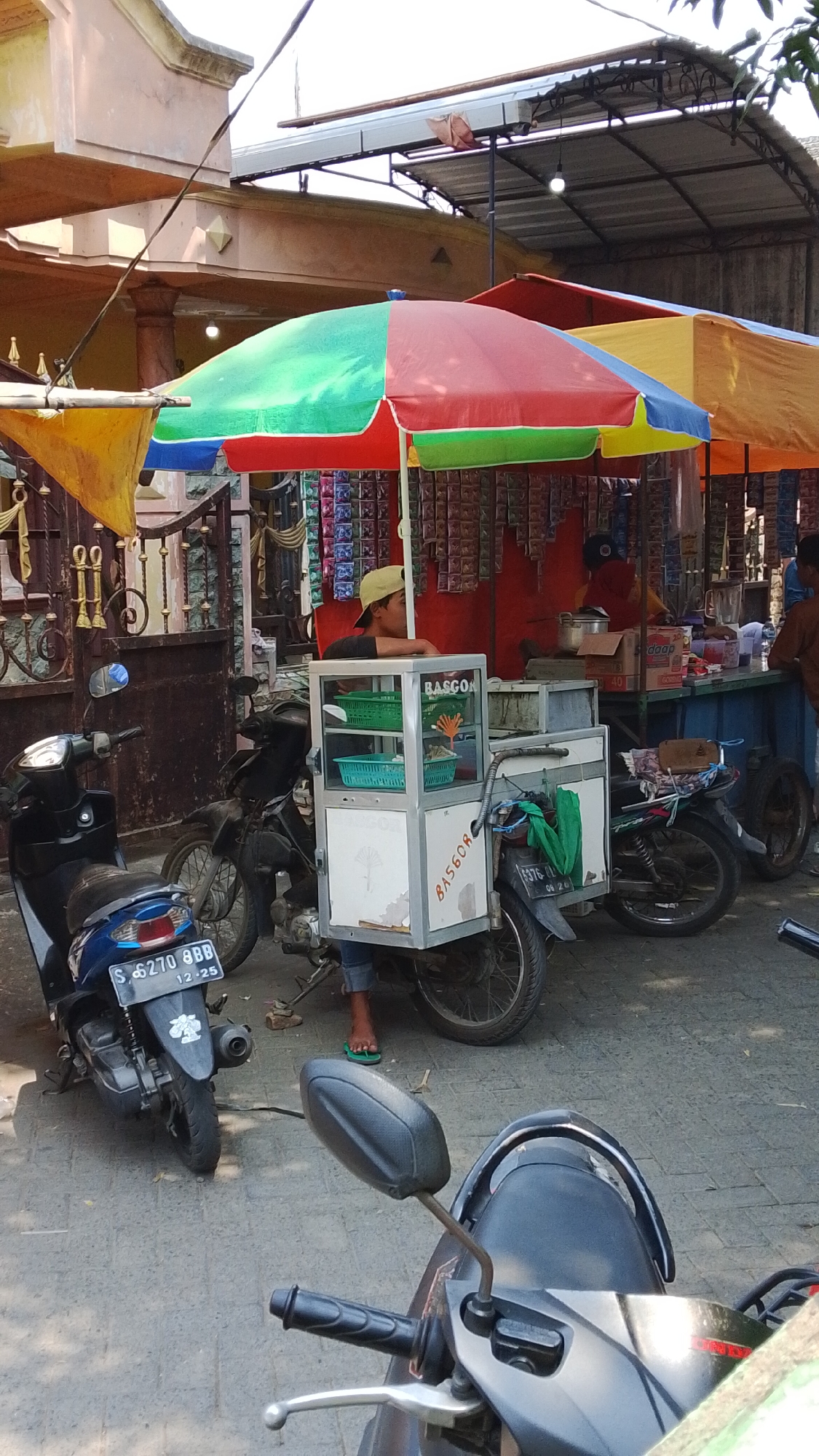 The image depicts a street scene in Mancar, Indonesia, featuring a food stall operated from a modified motorcycle. The stall is set up under a multi-colored umbrella, with red, green, blue, and yellow panels. In front of the stall, a young person wearing a beige cap and dark t-shirt stands behind a white food cart. The cart has transparent panels displaying green baskets and is labeled "BASGO". Two motorcycles are parked next to the food stall. One is a blue scooter with a license plate reading "S 6270 BBB". The other is a black motorcycle. In the background, there are various items for sale, including packaged goods hanging on display, and a table with bottles and other items. The environment suggests an outdoor market or roadside setting.