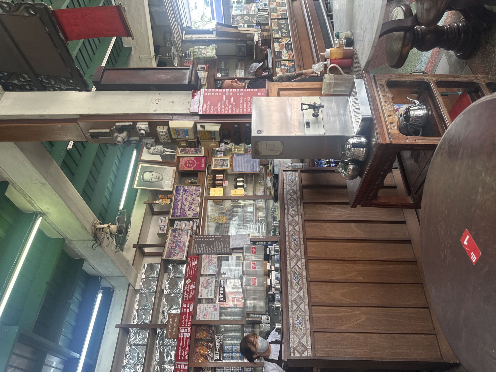 This image captures the interior of a traditional Thai cafe or eatery, likely located in Bangkok, characterized by its warm wooden paneling, exposed beams, and a mix of vintage and functional elements. The scene is observed from a slightly elevated, rotated perspective, looking across the main service area.
Two individuals are visible: one person wearing a cap and an arm tattoo is partially seen behind a counter or display shelf in the middle ground, reaching for something. Another person, wearing a face mask and a dark shirt over a white top, stands closer to the foreground, near a display of packaged goods.
The cafe's decor includes numerous framed photographs, some featuring groups of people, and what appear to be portraits, including one of an older man and another with the Chinese character "邢" (Xing). Overhead, an old-fashioned fan hangs from a beam, and fluorescent strip lights illuminate the space. A prominent red banner hangs from the ceiling.
Key objects include a large, stainless steel beverage dispenser, kettles, condiment bottles, and sturdy wooden tables and chairs, one with a red "L" sticker. Shelves display various products, including numerous foil-packaged items, likely coffee or snacks, and several clear dispensers.
Visible text, primarily in Thai, provides details about the cafe's offerings and service style. A large menu board lists drinks such as "ESPRESSO," "CAPPUCCINO," "MOCCA," "LATTE," "AMERICANO," "ICED COFFEE," "FRESH COFFEE," "MILK TEA," "BLACK TEA," "MILK," "OVALTINE, CACOA," "ICED LEMON TEA," and "YELLOW PLUM JUICE," along with their prices. Multiple signs read "SELF SERVICE บริการตัวเอง" (Self Service) and "สด 100%" (100% Fresh). Other texts include "น้ําสมุนไพร" (Herbal water/drink), "โคลัมโบ ศรีลังกาแฟสด" (Colombo Sri Lanka Fresh Coffee), and "เมล็ดกาแฟ munty" (Munty Coffee Beans). The overall impression is a bustling, established local establishment during daytime hours.